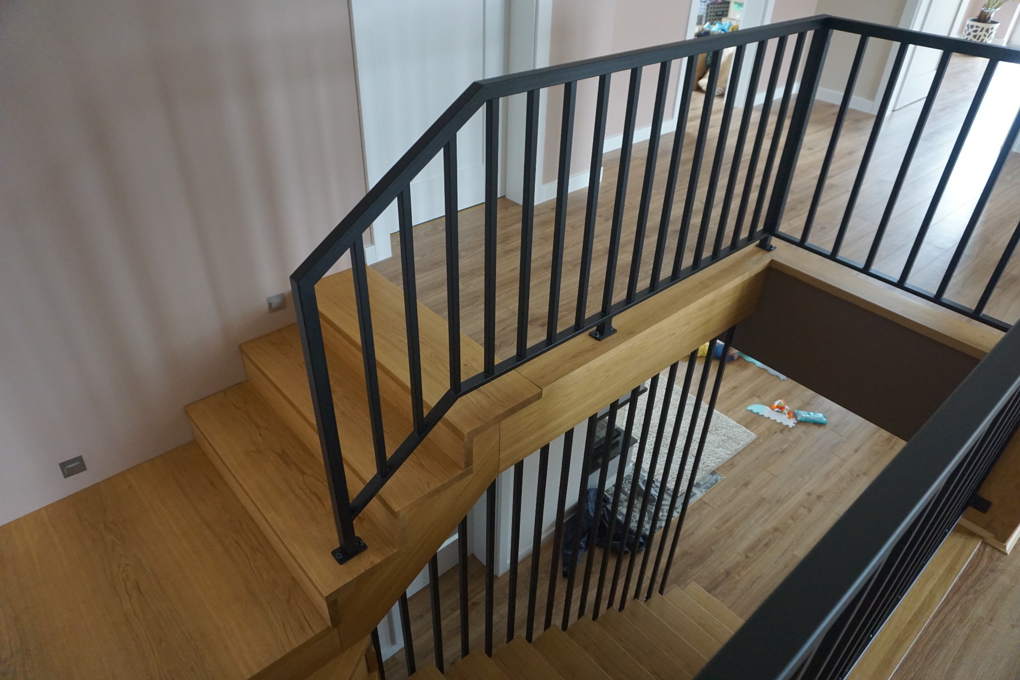 Kątowe ujęcie schodów z drewnianymi stopniami i czarną, metalową balustradą z pionowymi szczeblami. Widoczne jasne, drewniane stopnie i podłoga oraz zabawki na podłodze poniżej.
