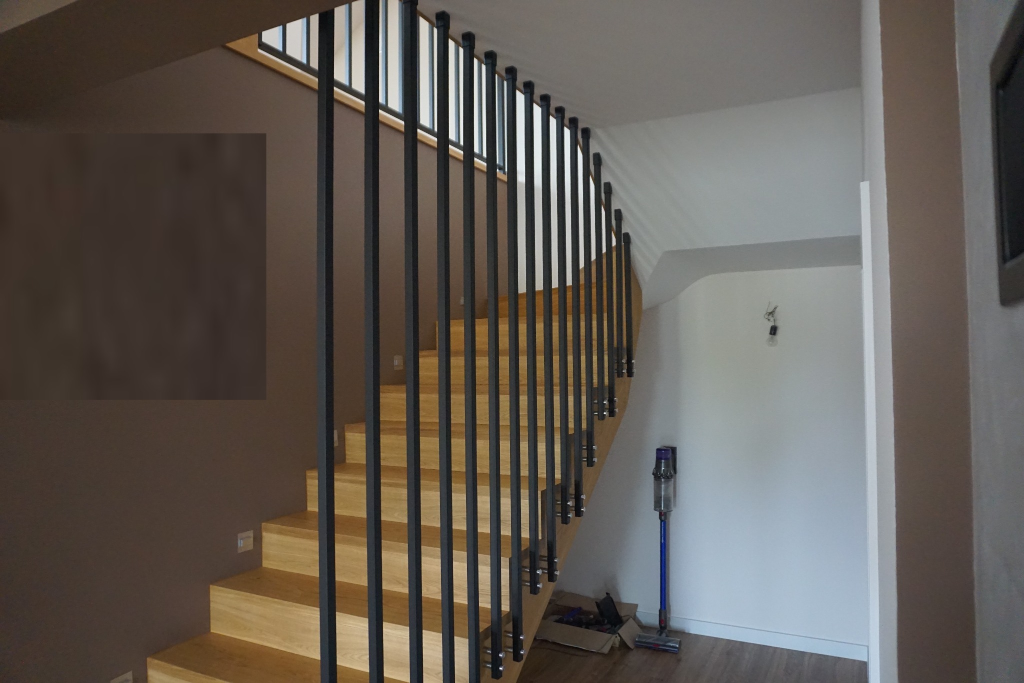 Drewniane schody z czarnymi, pionowymi balustradami metalowymi, widoczne z boku, z odkurzaczem stojącym w tle.