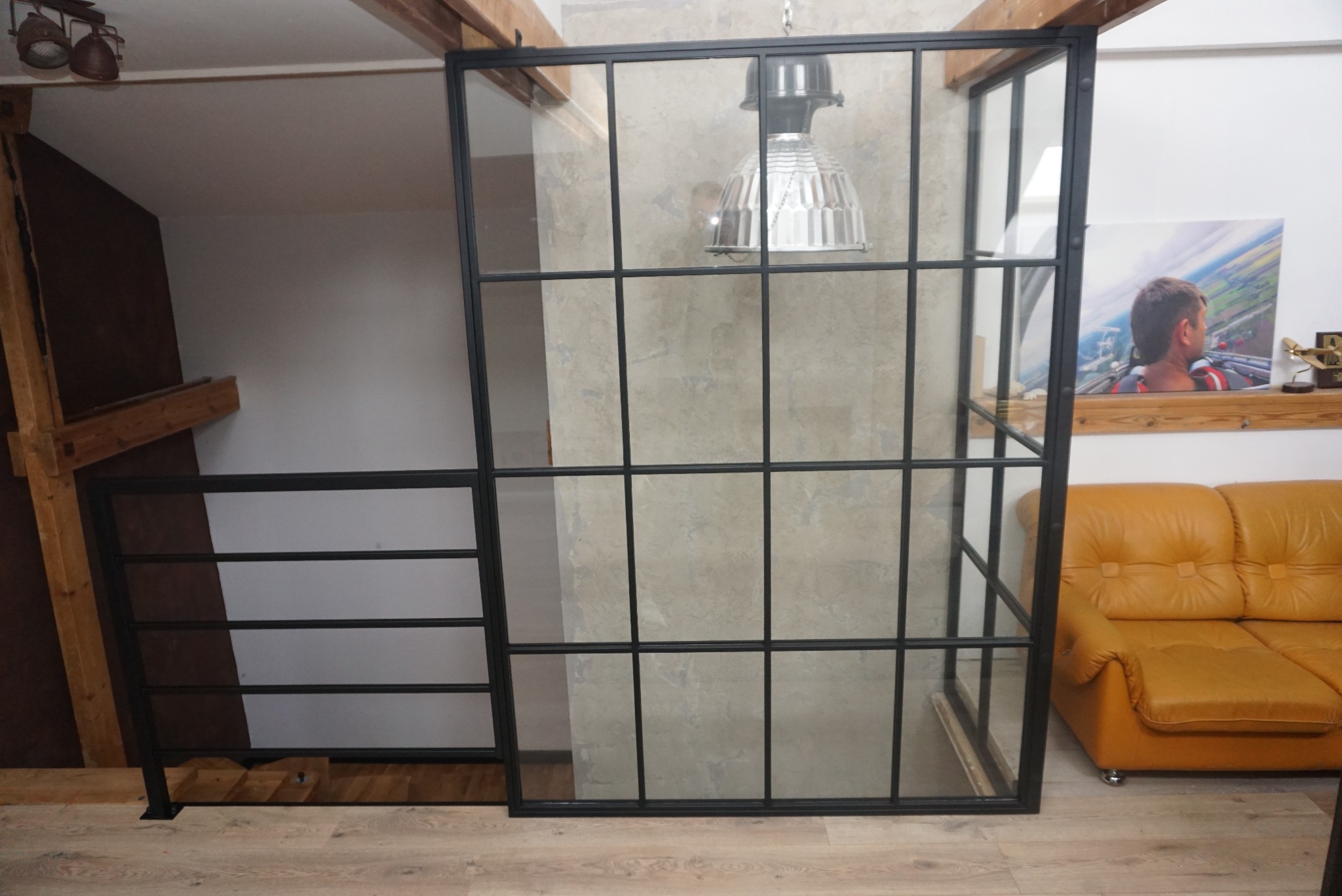 Nowoczesna balustrada schodowa z czarnych metalowych profili i szkła, oddzielająca przestrzeń schodów od salonu z żółtą sofą, widoczna lampa w stylu industrialnym i drewniane elementy konstrukcyjne.