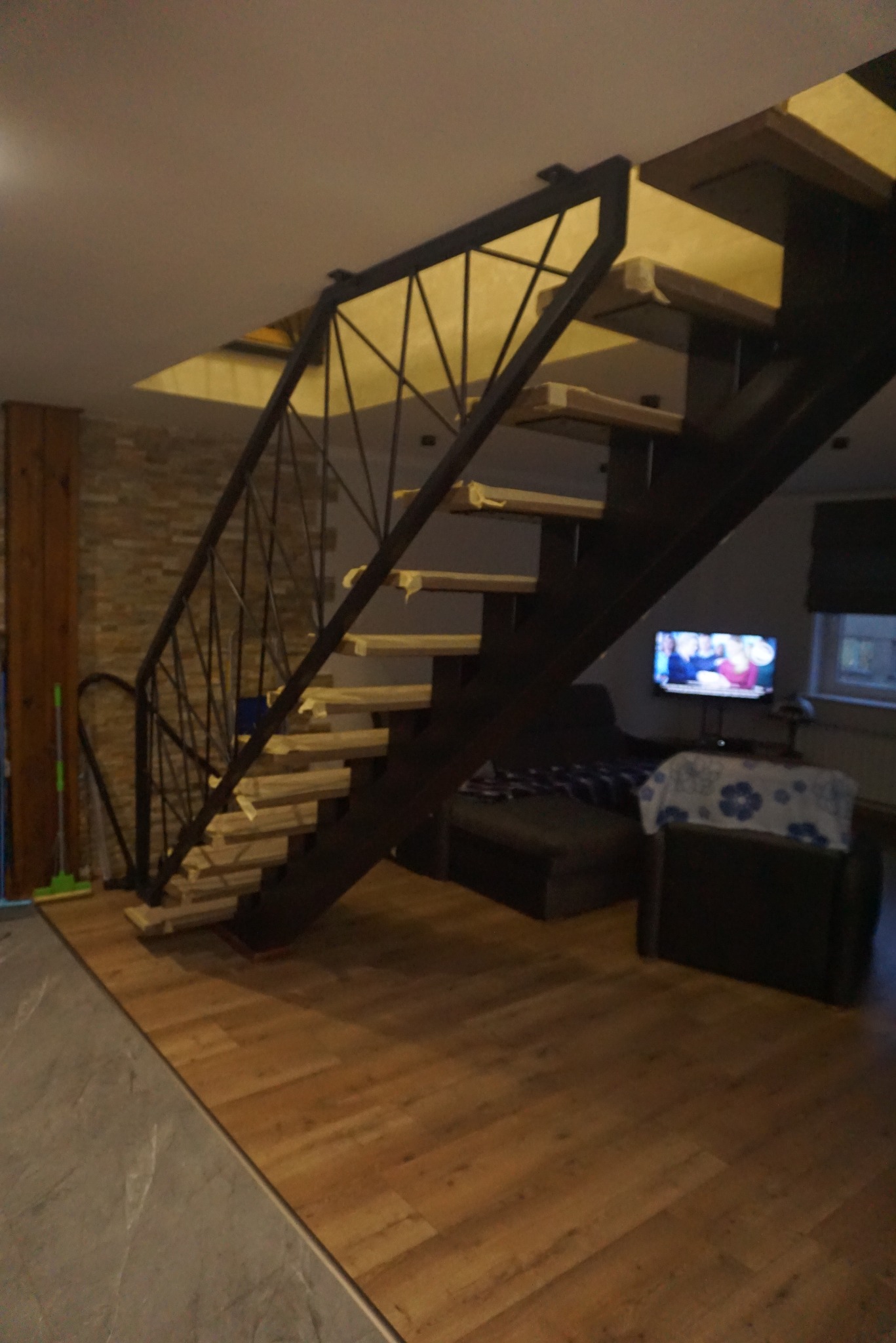 Nowoczesne schody wewnętrzne z czarną metalową konstrukcją i drewnianymi stopniami, widok na salon z telewizorem w tle.