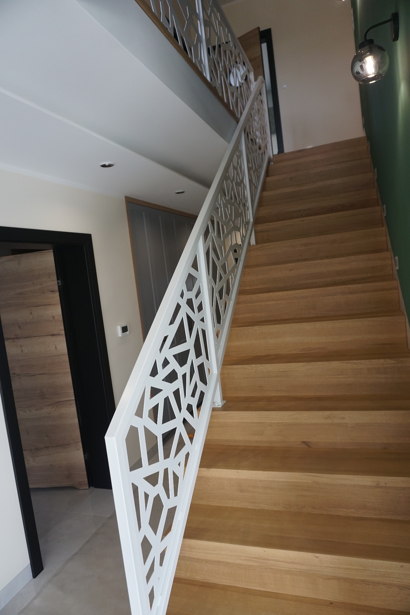 Klatka schodowa z drewnianymi stopniami i białą, ażurową balustradą z geometrycznym wzorem, widziana z dołu, z lampą ścienną w tle.
