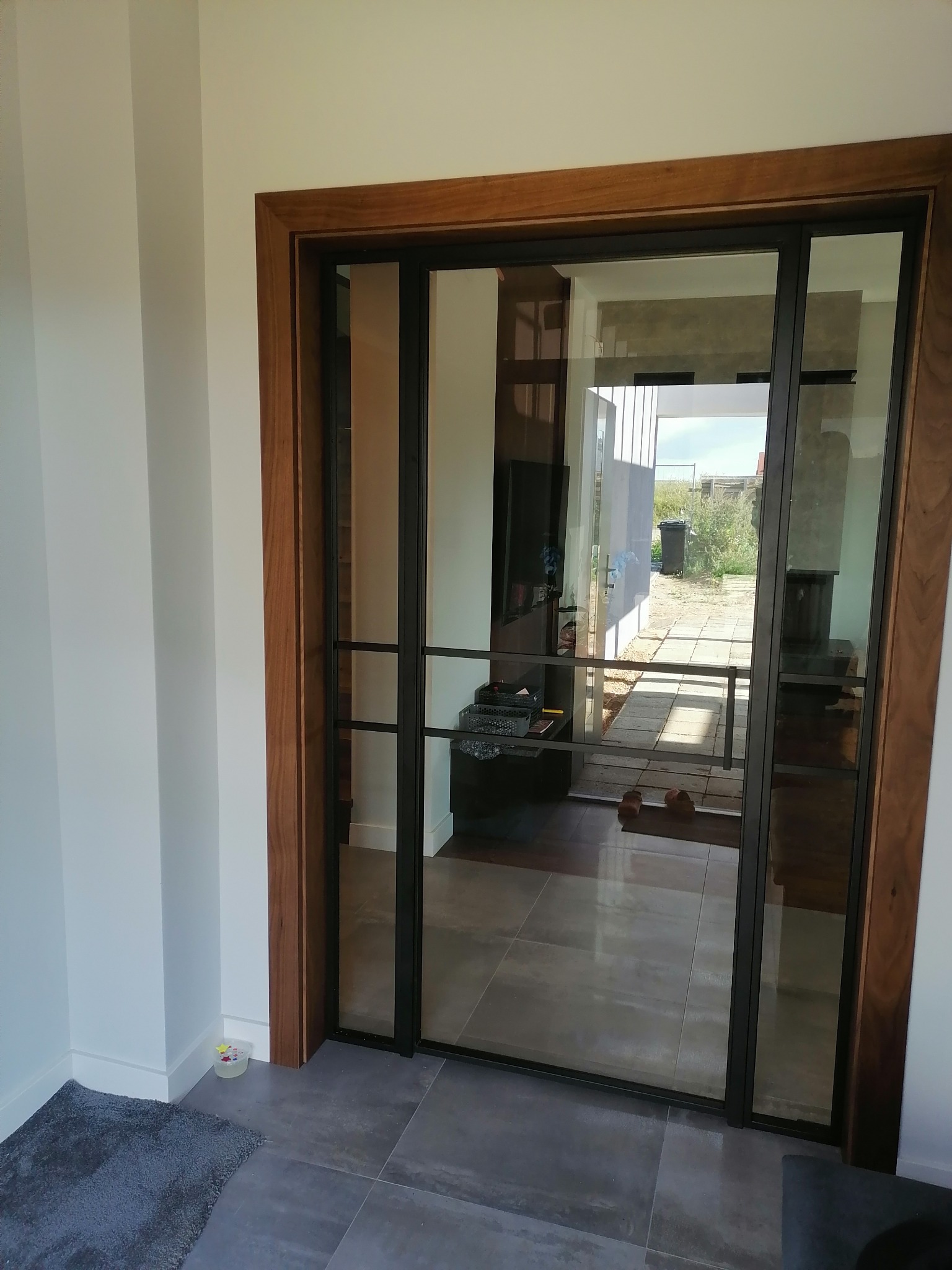 Przeszklone drzwi przesuwne z czarną metalową ramą i drewnianą ościeżnicą, widok na taras i ogród.