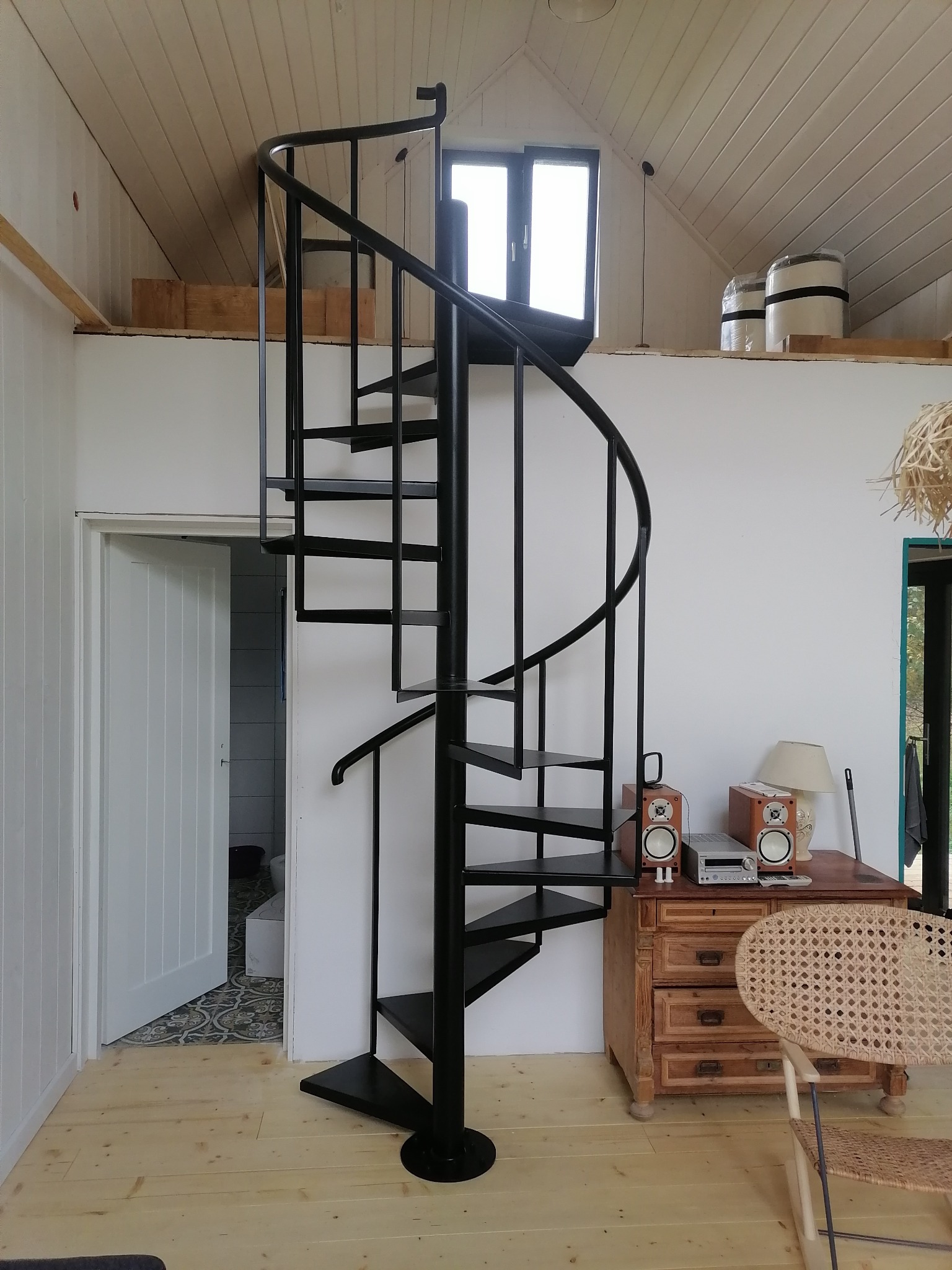 Czarne, spiralne schody w jasnym pomieszczeniu z drewnianą podłogą, widoczne elementy wystroju wnętrza: komoda, głośniki i otwarte drzwi do łazienki.
