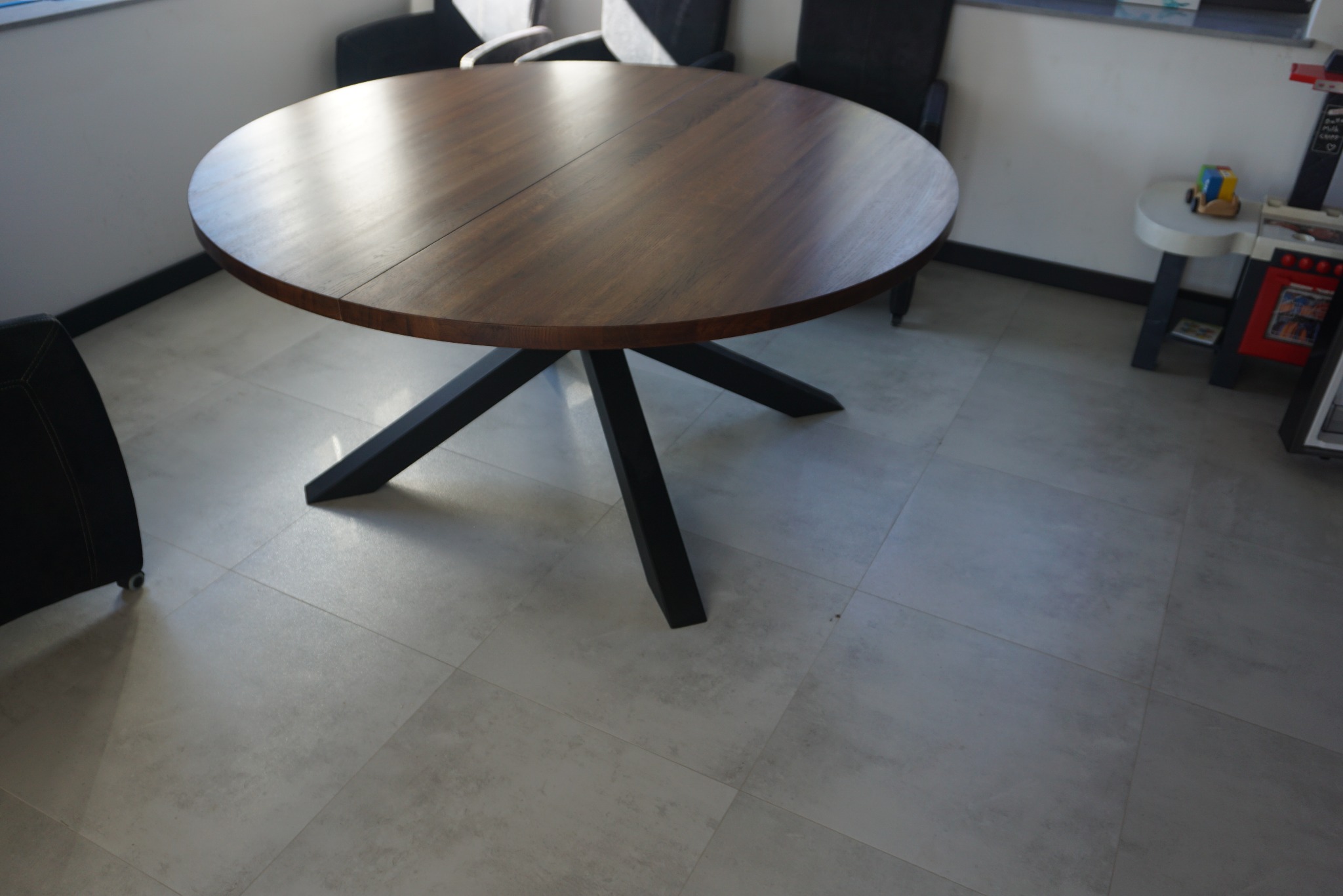 Okrągły stół z ciemnego drewna na czarnej, metalowej podstawie w nowoczesnym wnętrzu, widoczne krzesła i fragment podłogi z szarych płytek.