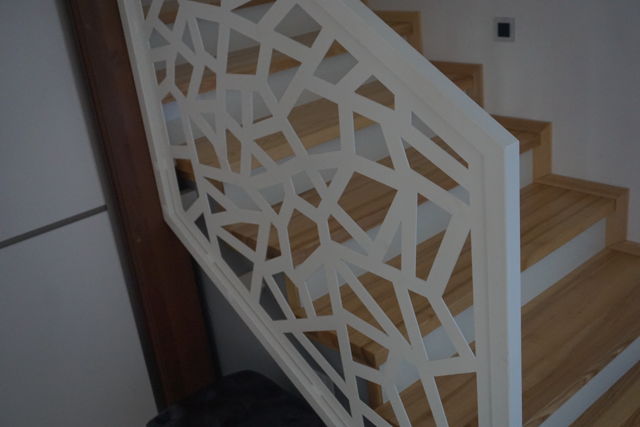 Biała, ażurowa balustrada schodowa z geometrycznym wzorem, drewniane schody w tle, minimalistyczny design.