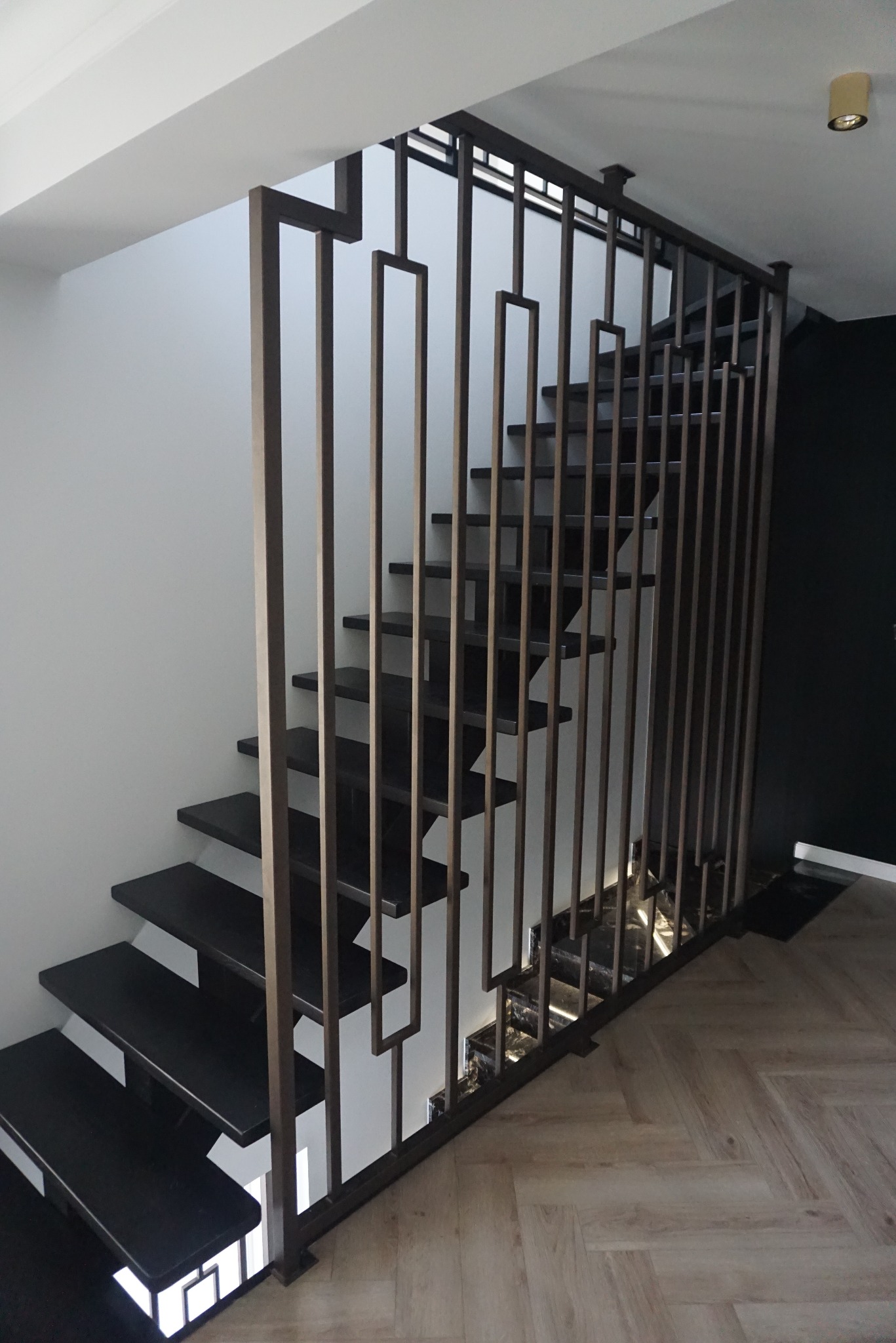 Nowoczesne schody z czarnymi stopniami i metalową balustradą z prostokątnymi elementami, oświetlenie LED pod schodami, parkiet ułożony w jodełkę.