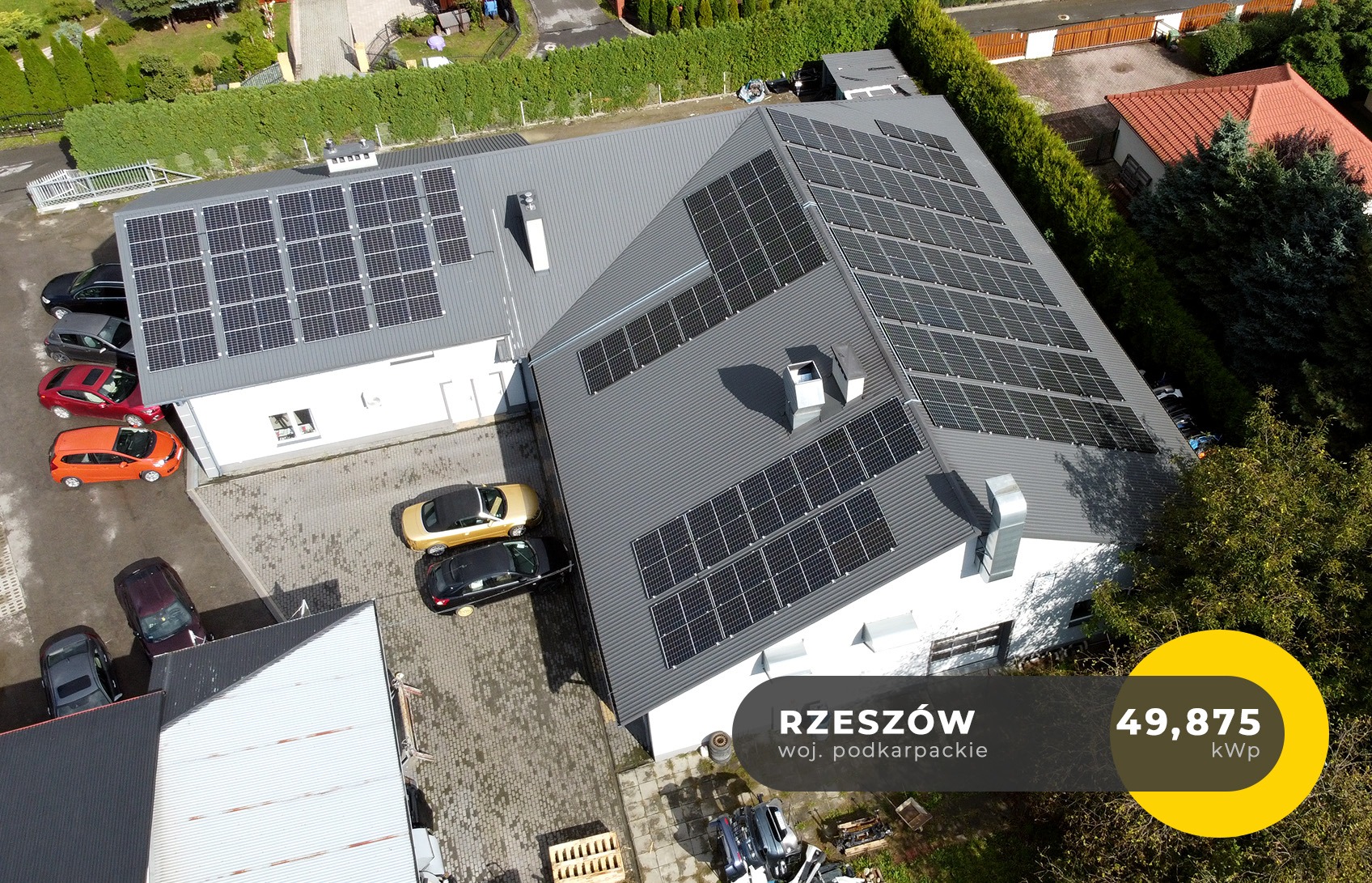 Widok z góry na budynek z szarym dachem pokrytym panelami słonecznymi, zaparkowane samochody w różnych kolorach, w tle zieleń drzew i krzewów, w prawym dolnym rogu żółty element graficzny z napisem...