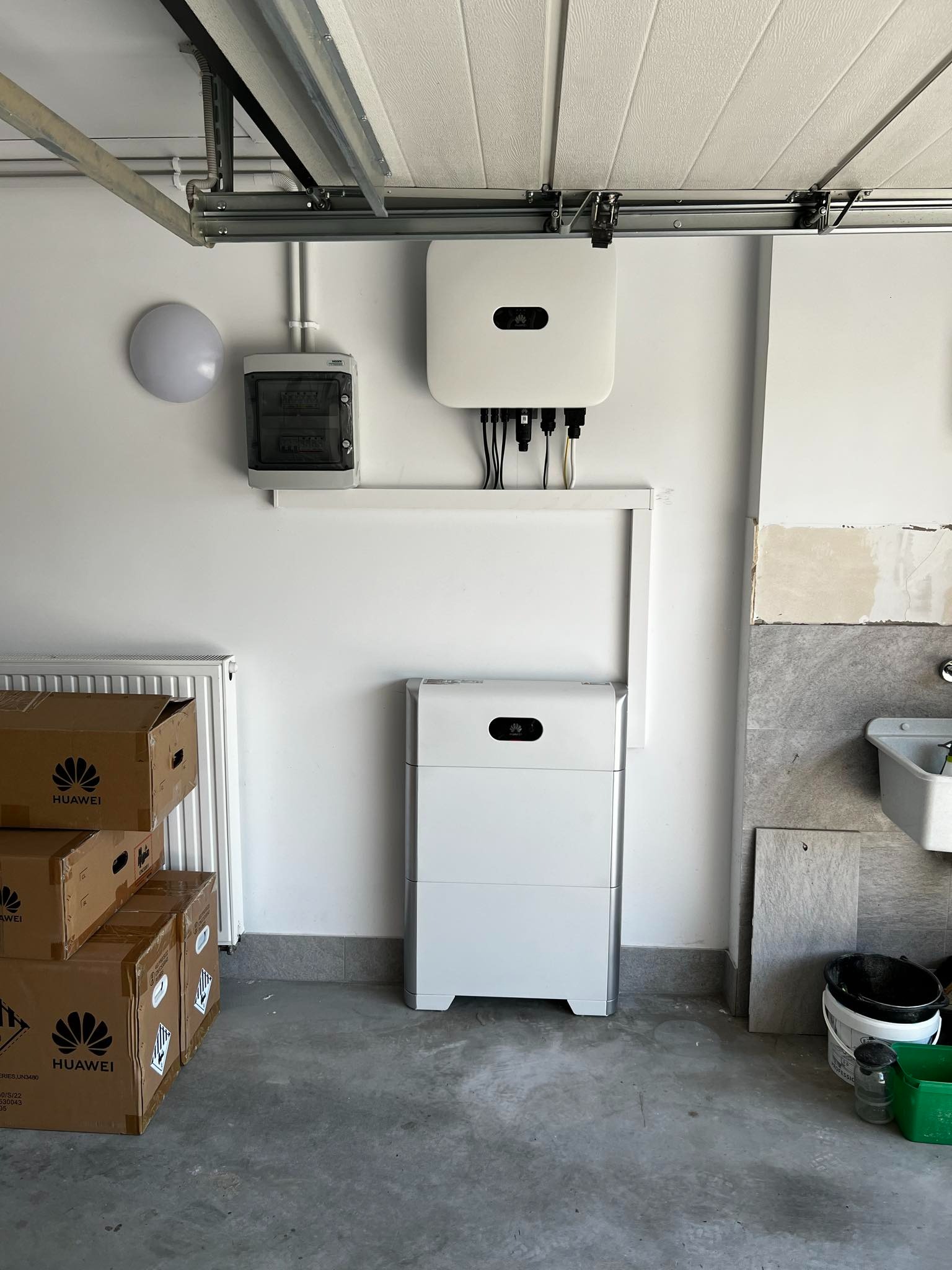Wnętrze garażu z zamontowanym magazynem energii Huawei, inwerterem i skrzynką elektryczną na białej ścianie, obok sterta kartonów z logo Huawei i umywalka z wiadrami.