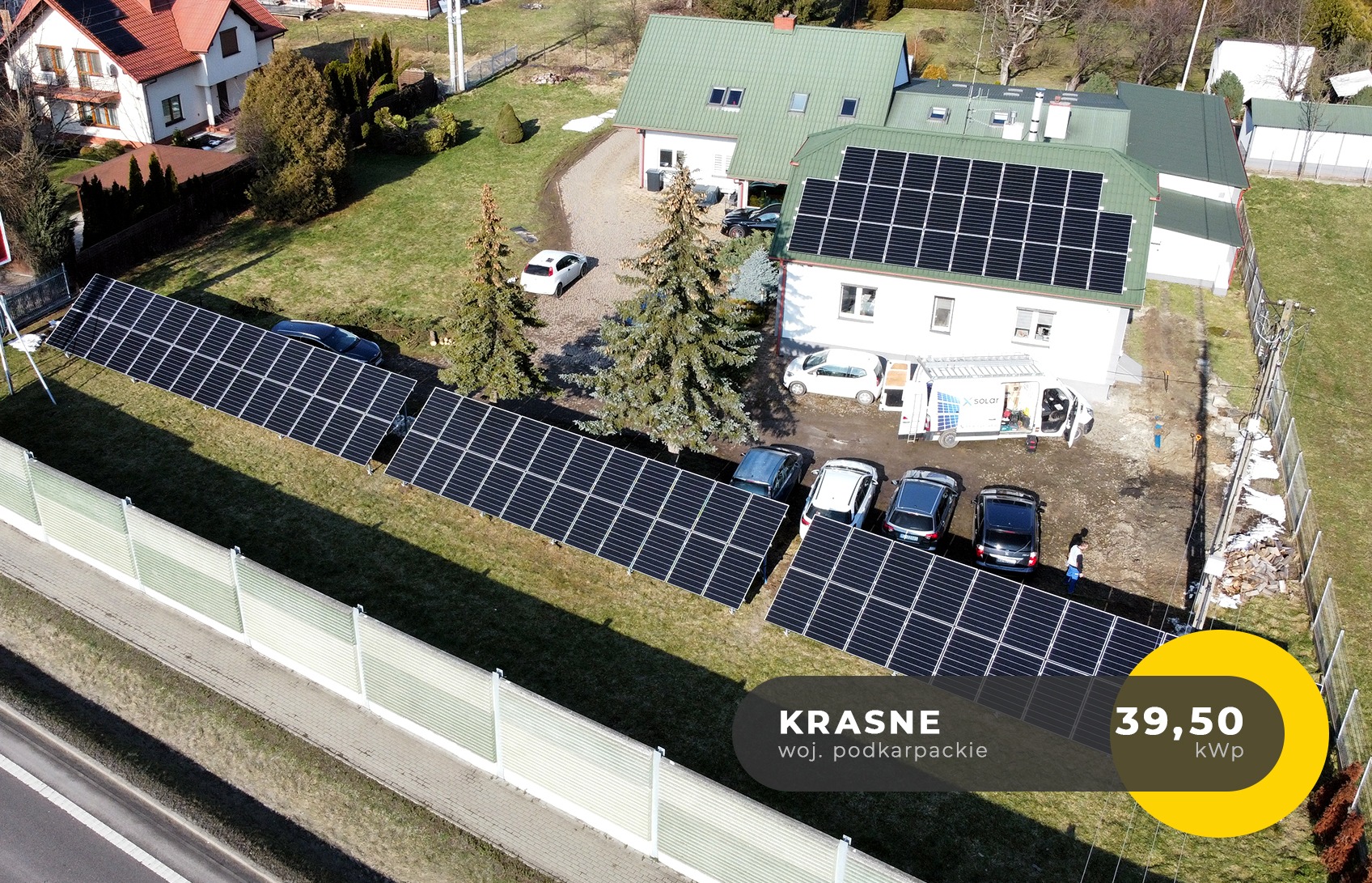 Widok z lotu ptaka na instalację paneli słonecznych na gruncie obok domu z panelami słonecznymi na dachu, kilka samochodów zaparkowanych obok białego busa z logo firmy instalacyjnej, miejscowość...