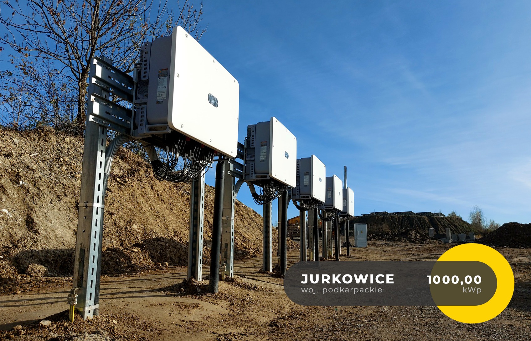 Rząd białych falowników zamontowanych na metalowych konstrukcjach na tle niebieskiego nieba i ziemi, widoczne kable i elementy montażowe.