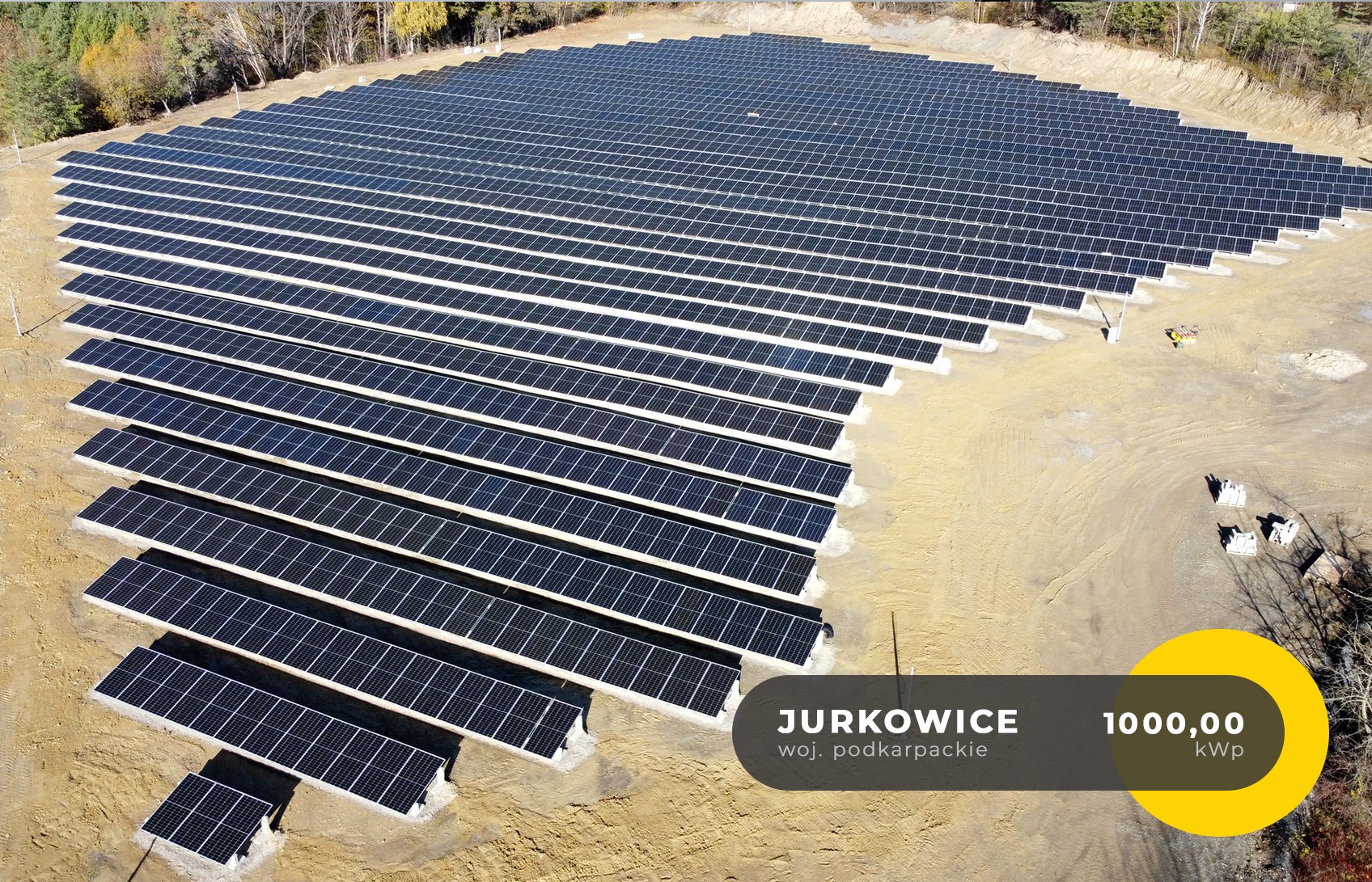 Rozległa farma paneli słonecznych w Jurkowicach, województwo podkarpackie, z żółtym oznaczeniem 1000,00 kWp, widok z lotu ptaka na rzędy paneli na piaszczystym terenie, zalesione wzgórze w tle.
