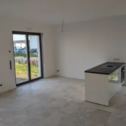 Surowe wnętrze z jasnymi ścianami, płytkami podłogowymi imitującymi kamień i białą wyspą kuchenną z czarnym blatem, widok na zewnątrz przez przesuwne drzwi balkonowe.