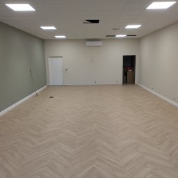 Cezary krawczyk - Jasne panele ułożone w jodełkę w przestronnym, odnowionym wnętrzu z białymi ścianami i sufitem kasetonowym. Widoczny klimatyzator i drzwi.