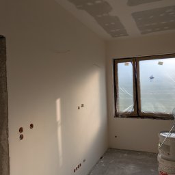 Cezary krawczyk - Wnętrze w trakcie prac wykończeniowych: gładkie, białe ściany z widocznymi otworami na gniazdka, okno zabezpieczone folią, ślady gładzi na suficie i podłodze, wiaderka z farbą.