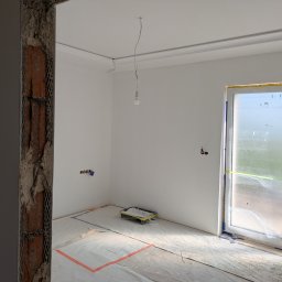 Cezary krawczyk - Wnętrze pokoju po szpachlowaniu ścian, przygotowane do malowania. Widoczne zabezpieczenia folią, listwy sufitowe i wisząca żarówka. Surowy stan wykończenia.