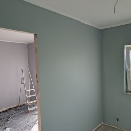 Cezary krawczyk - Świeżo pomalowane na gładko ściany w odcieniach szarości i błękitu, z widoczną drabiną i zabezpieczoną podłogą, ukazujące postęp prac wykończeniowych.