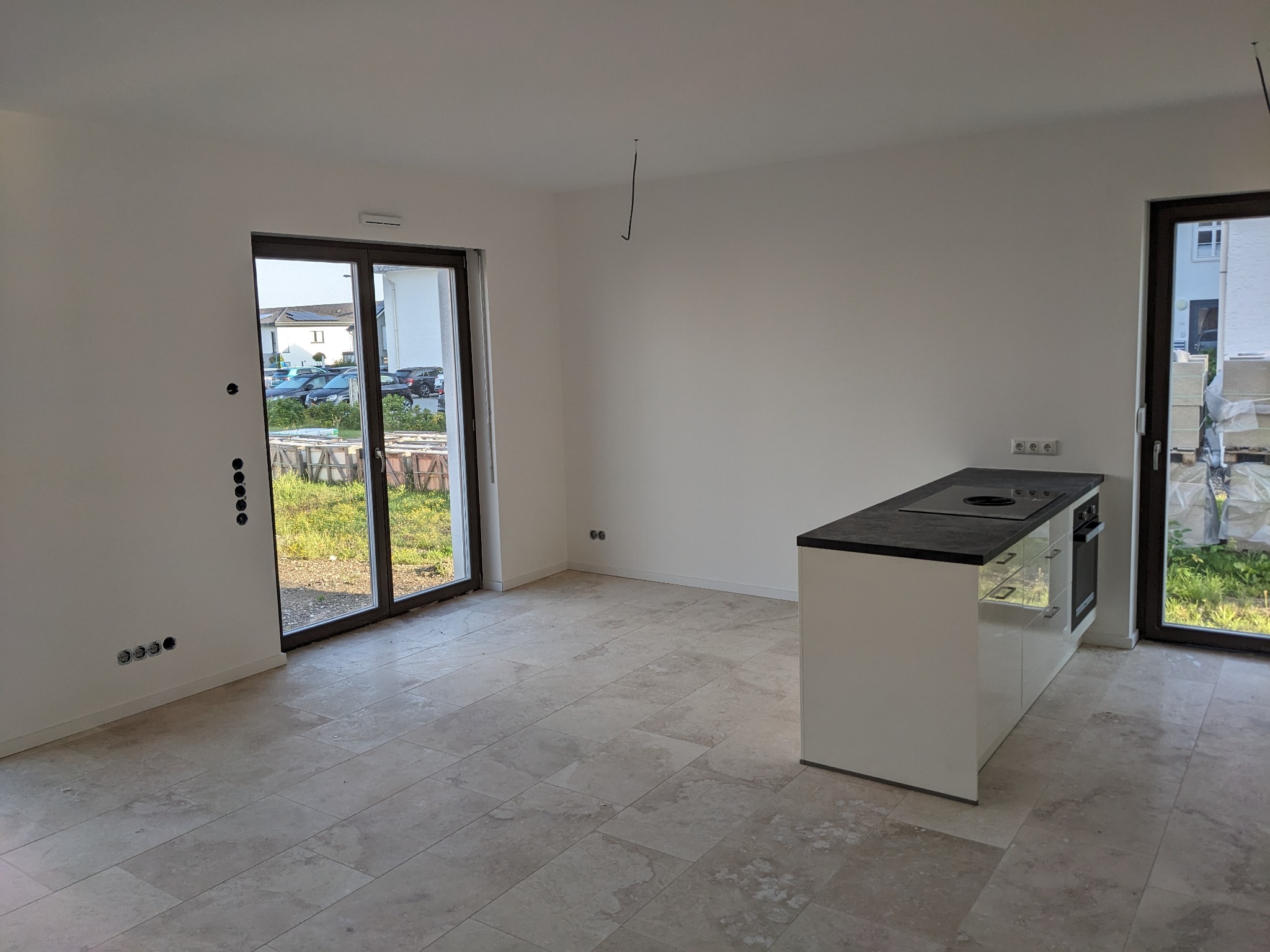 Surowe wnętrze z jasnymi ścianami, płytkami podłogowymi imitującymi kamień i białą wyspą kuchenną z czarnym blatem, widok na zewnątrz przez przesuwne drzwi balkonowe.