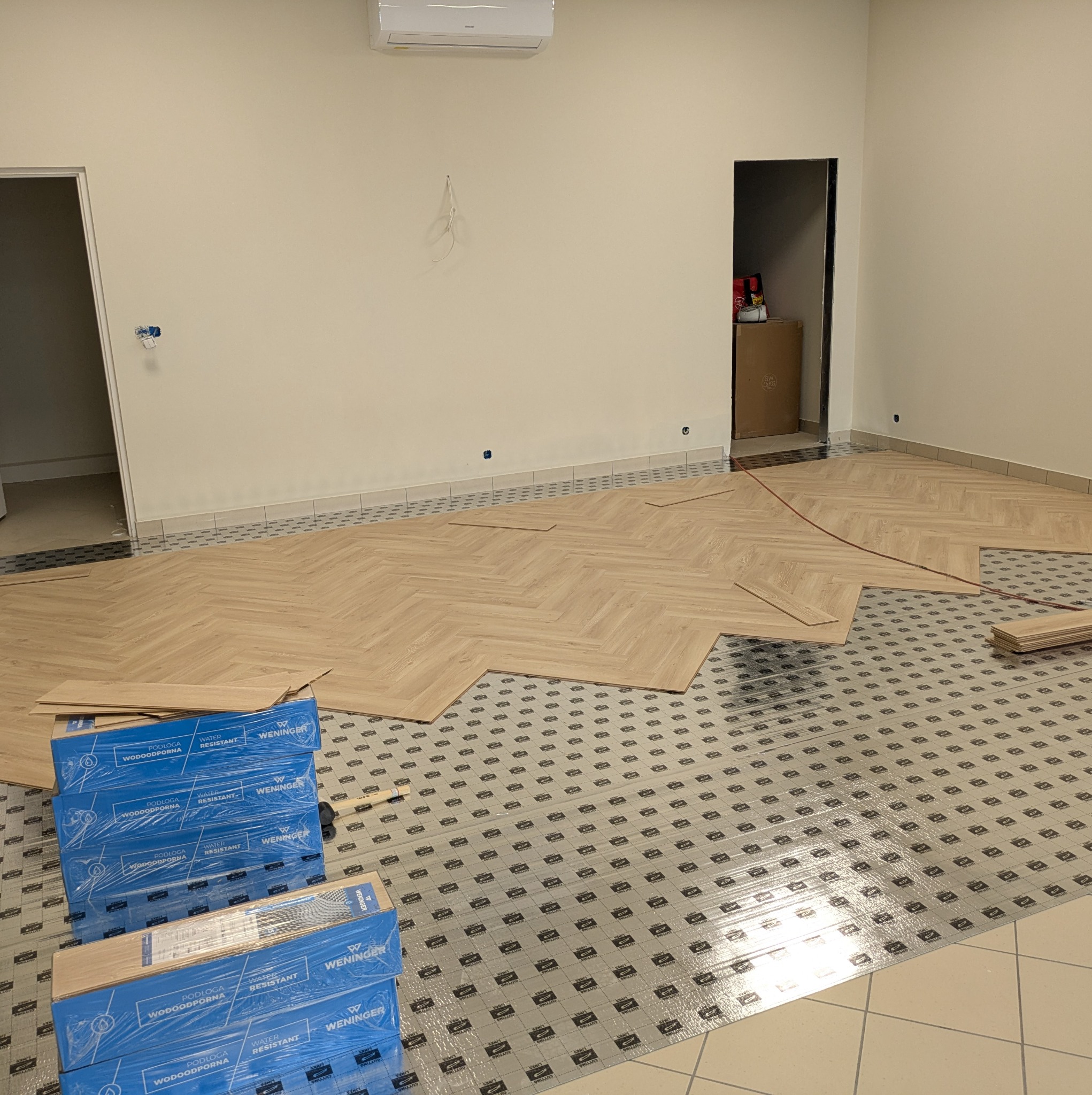 Układanie paneli w jodełkę na podkładzie. Częściowo ułożona podłoga, paczki paneli Wenninger. Widoczne kable wystające ze ściany i klimatyzator.