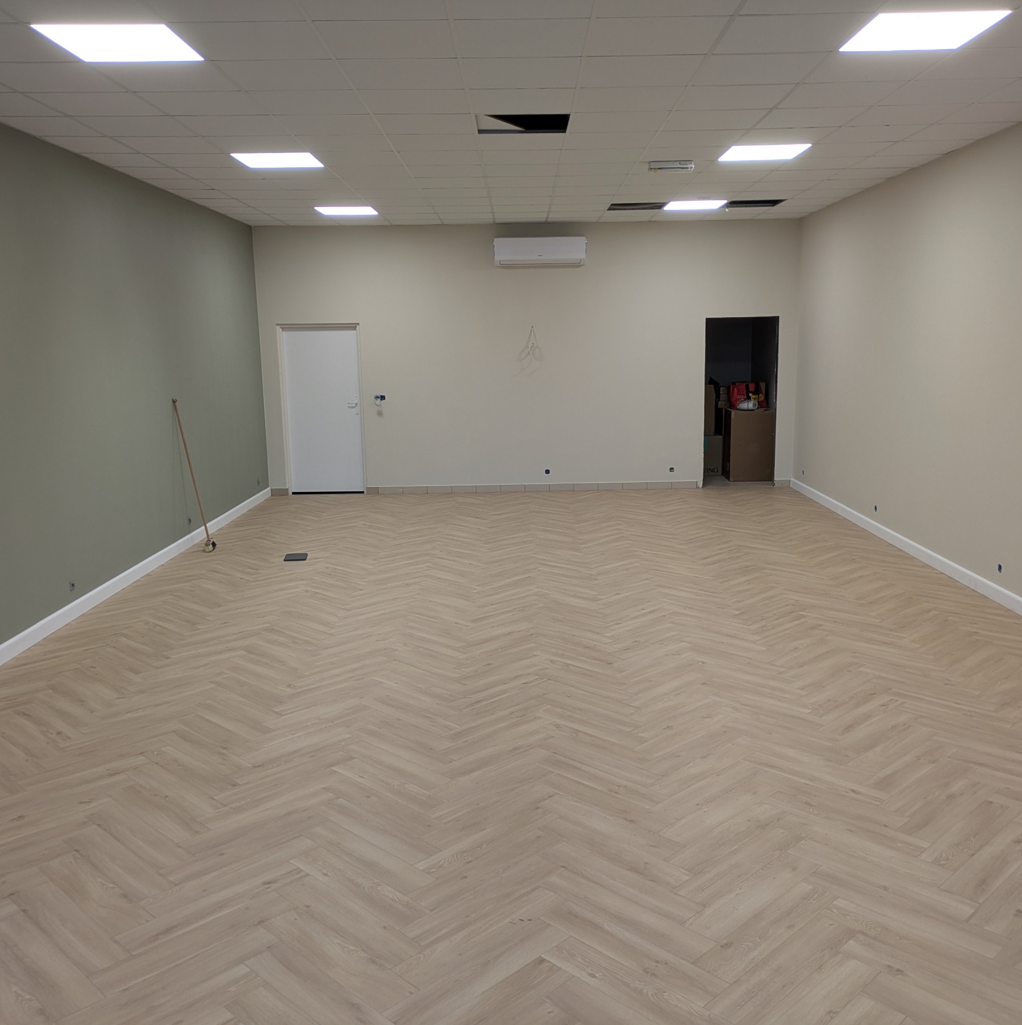 Jasne panele ułożone w jodełkę w przestronnym, odnowionym wnętrzu z białymi ścianami i sufitem kasetonowym. Widoczny klimatyzator i drzwi.