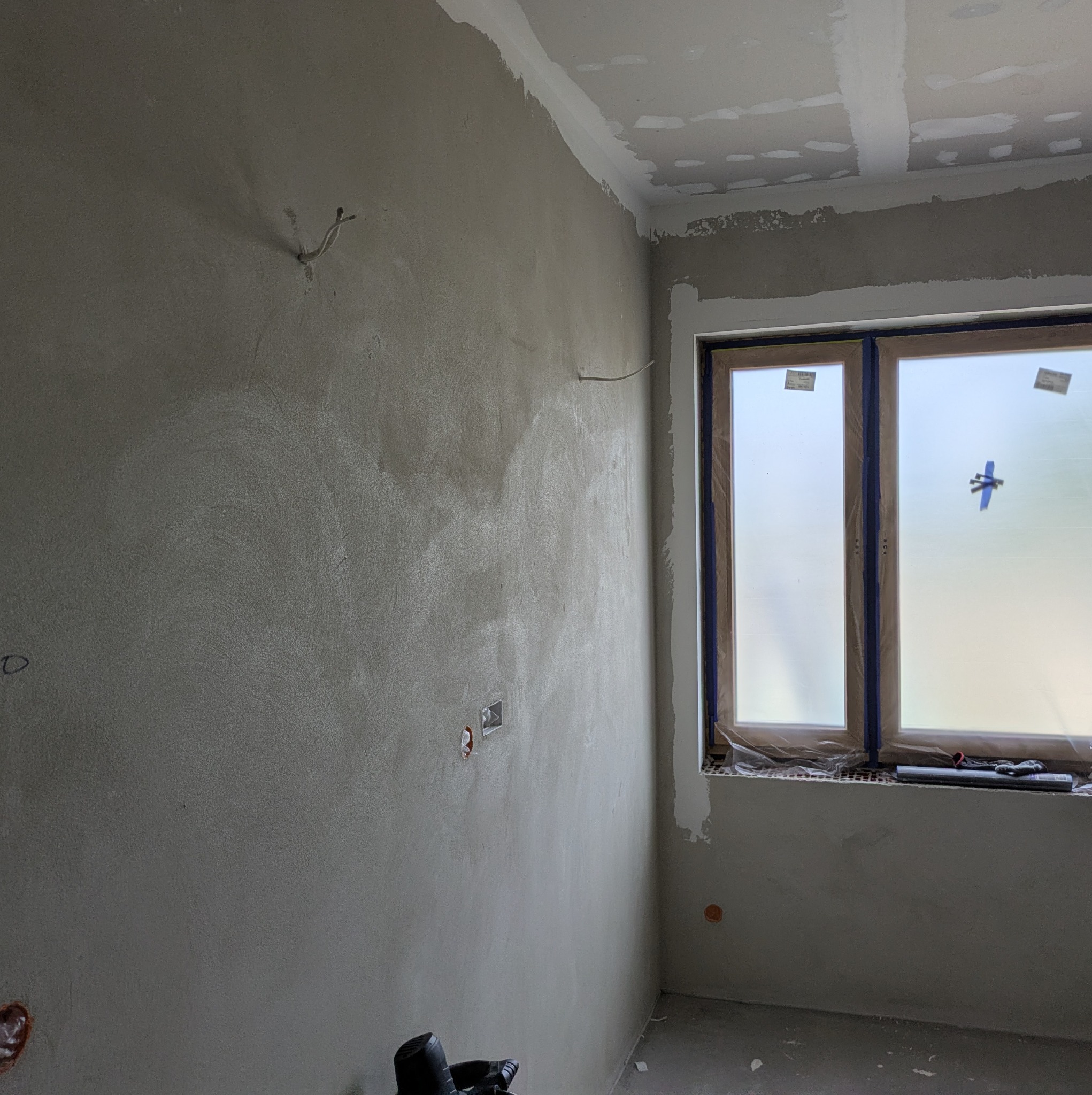 Ściana po szpachlowaniu z widocznymi śladami po narzędziach, przygotowana do dalszych prac wykończeniowych. Okno zabezpieczone folią malarską.