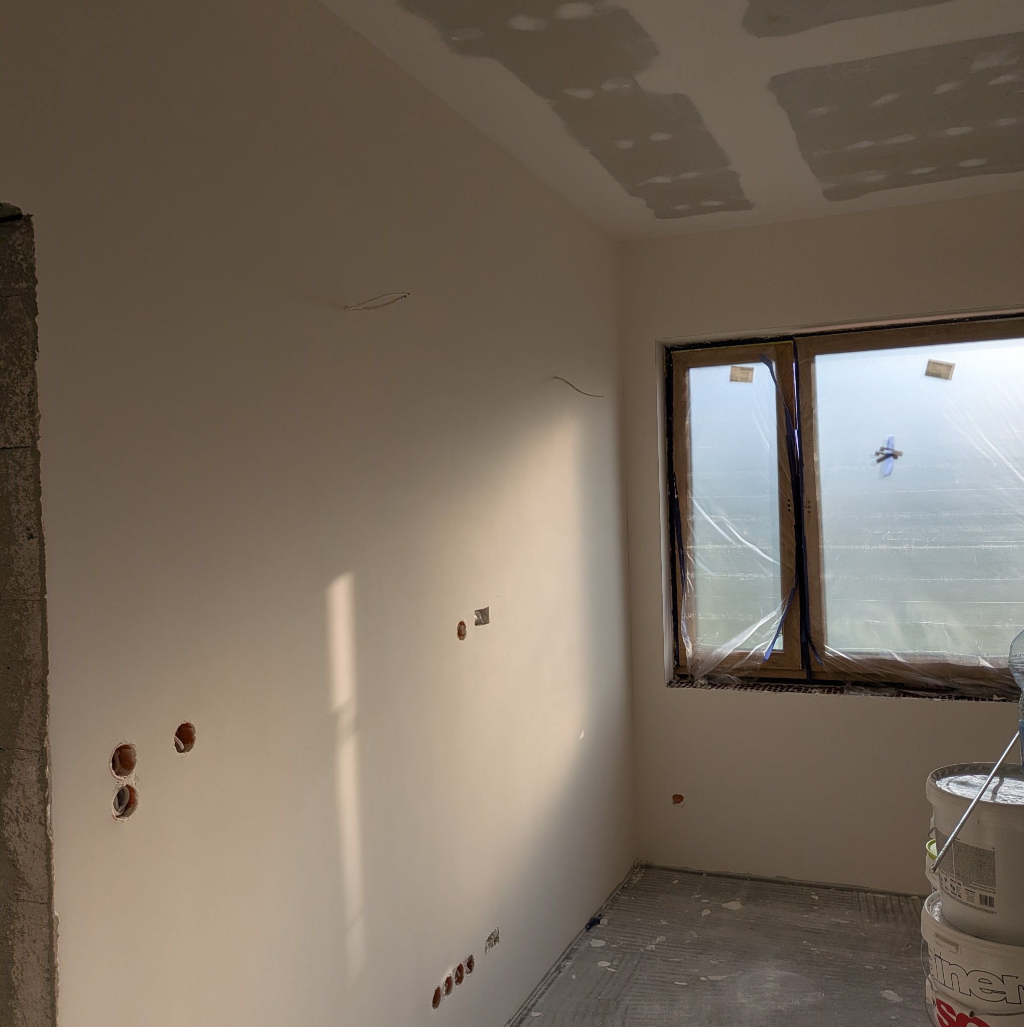 Wnętrze w trakcie prac wykończeniowych: gładkie, białe ściany z widocznymi otworami na gniazdka, okno zabezpieczone folią, ślady gładzi na suficie i podłodze, wiaderka z farbą.