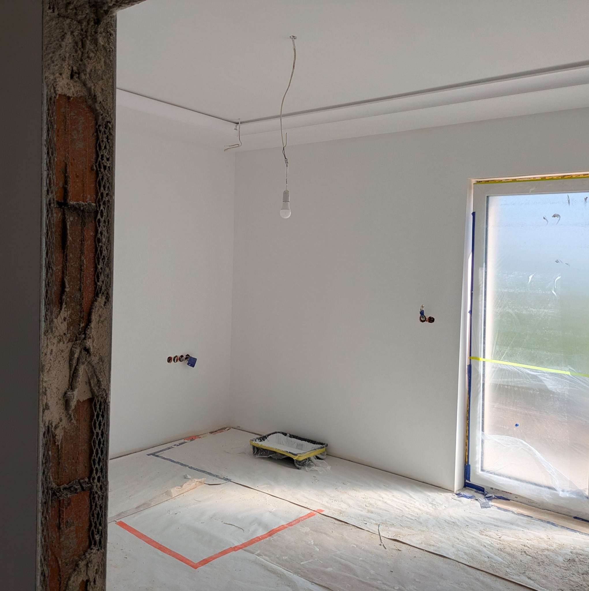Wnętrze pokoju po szpachlowaniu ścian, przygotowane do malowania. Widoczne zabezpieczenia folią, listwy sufitowe i wisząca żarówka. Surowy stan wykończenia.