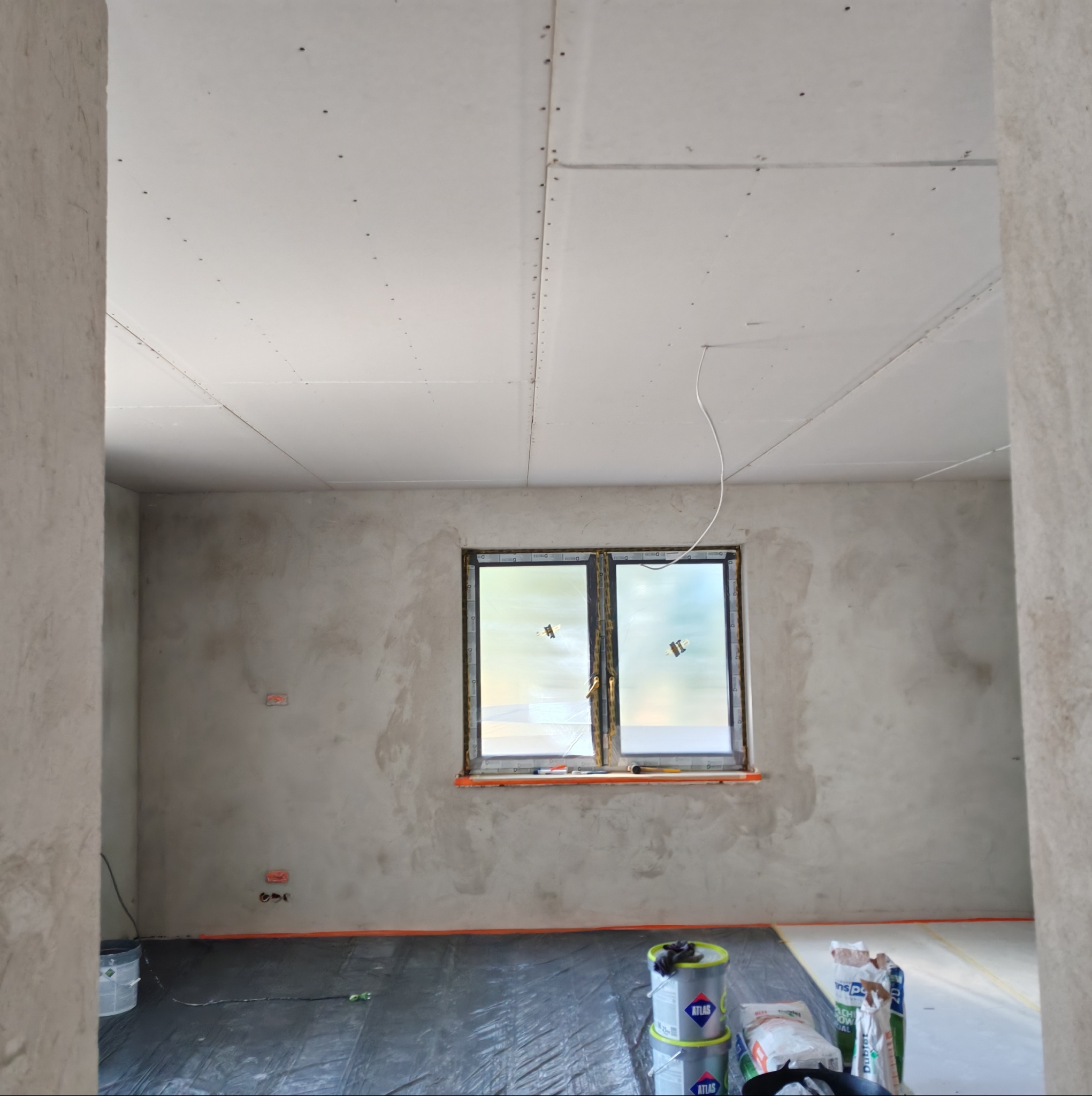 Pokój w trakcie prac wykończeniowych: ściany po szpachlowaniu, sufit z płyt karton-gips, okno z folią ochronną, podłoga zabezpieczona folią, widoczne materiały budowlane.