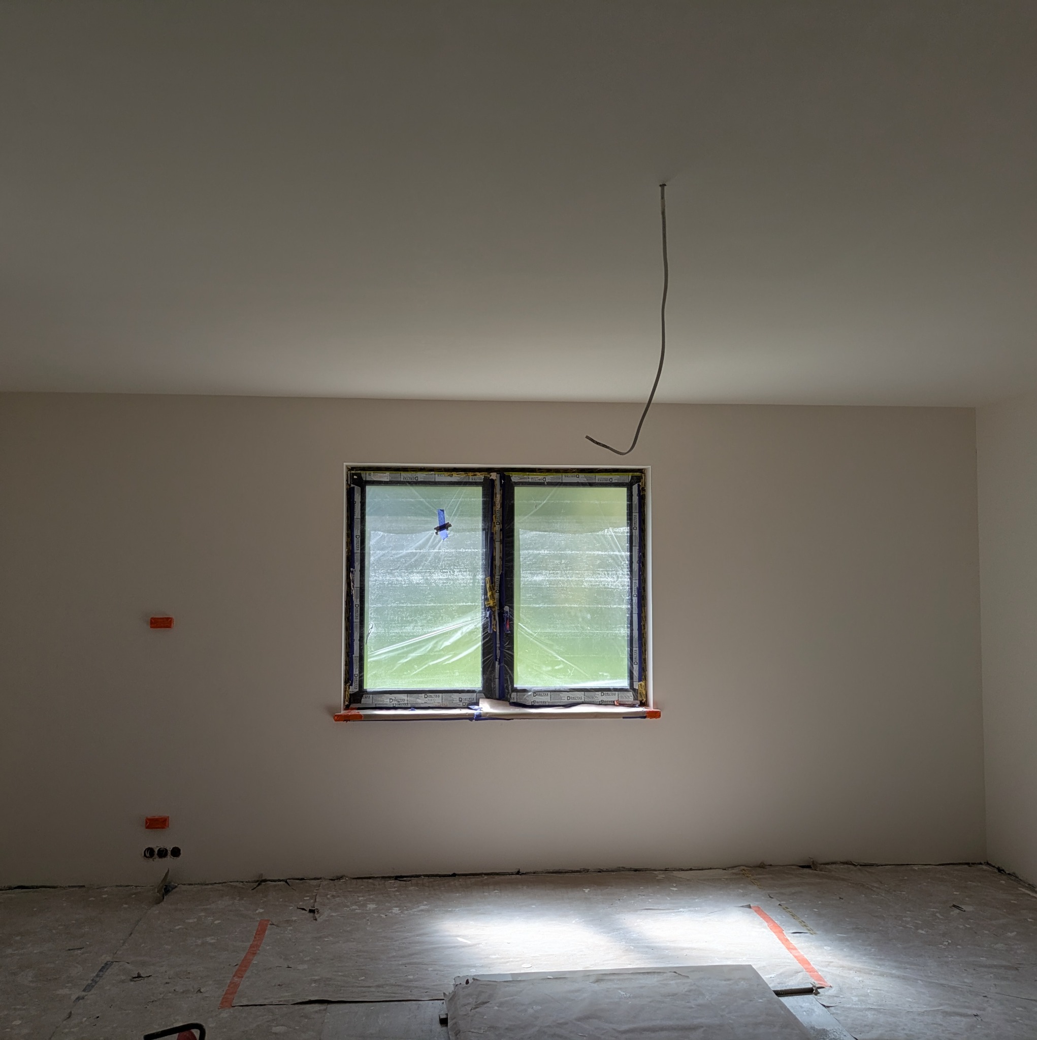 Pokój po szpachlowaniu, z zabezpieczonym oknem folią. Widoczne puszki elektryczne i przewód zwisający z sufitu. Podłoga zabezpieczona folią malarską.