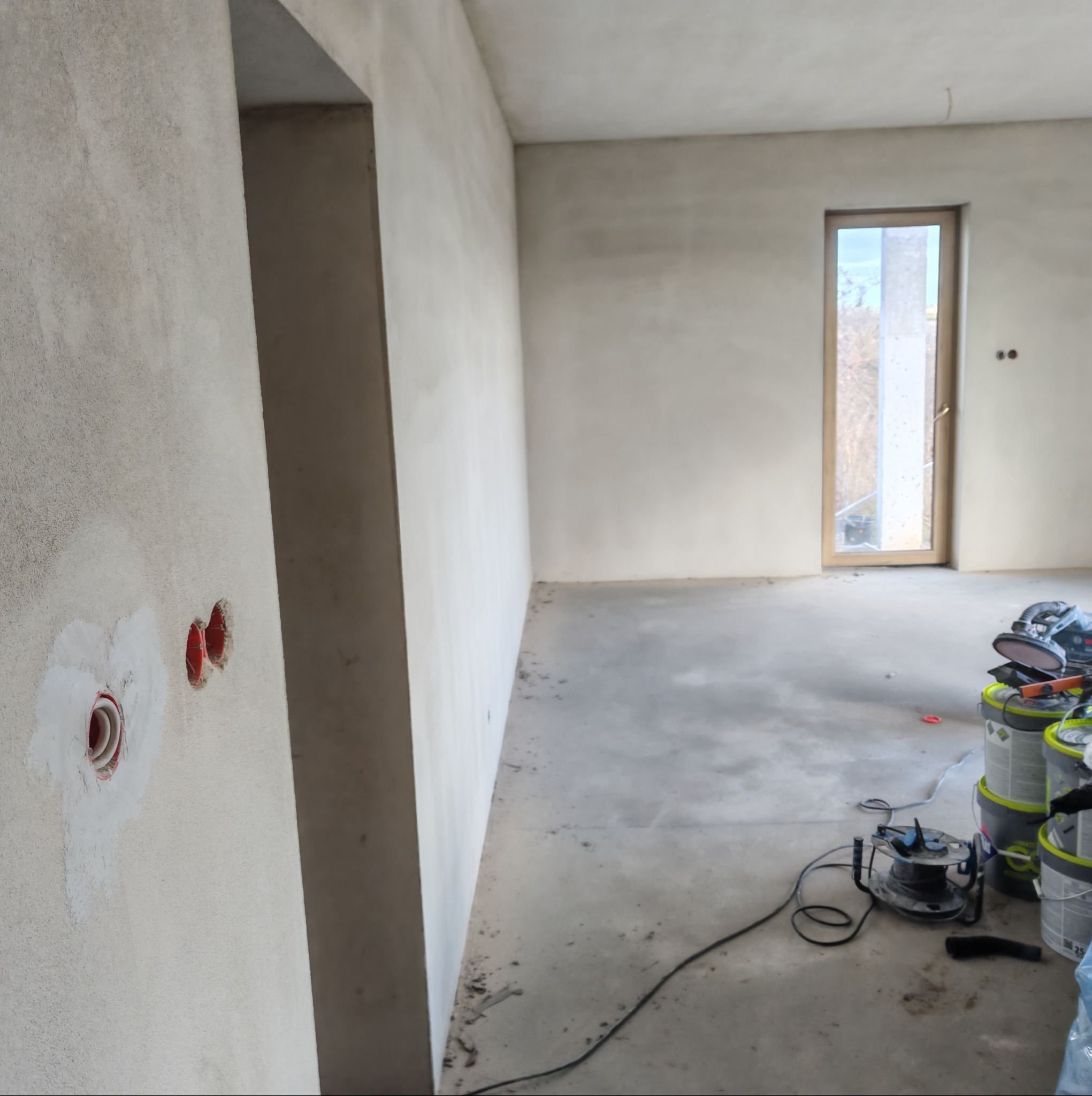 Wnętrze w trakcie prac wykończeniowych: ściany po szpachlowaniu, widoczne puszki elektryczne, podłoga surowa, w tle okno i narzędzia malarskie.