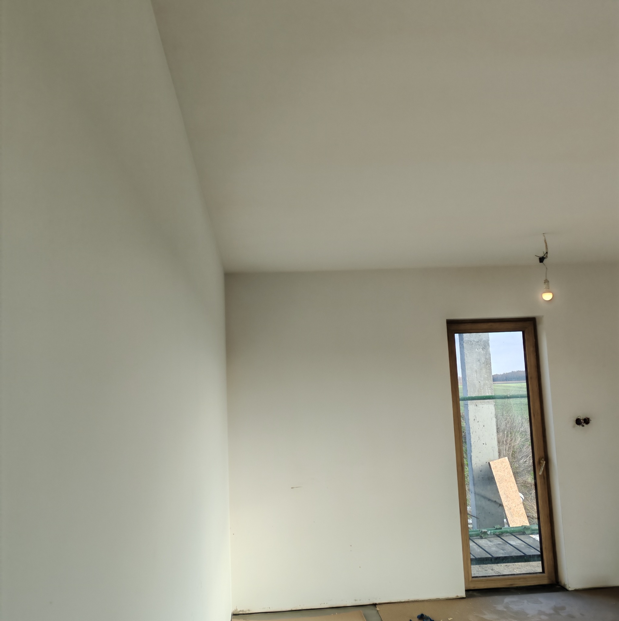 Wnętrze pokoju po szpachlowaniu ścian na biało, widoczne gładkie powierzchnie, okno z widokiem na zewnątrz i wystające kable z żarówką. Minimalistyczny kadr.