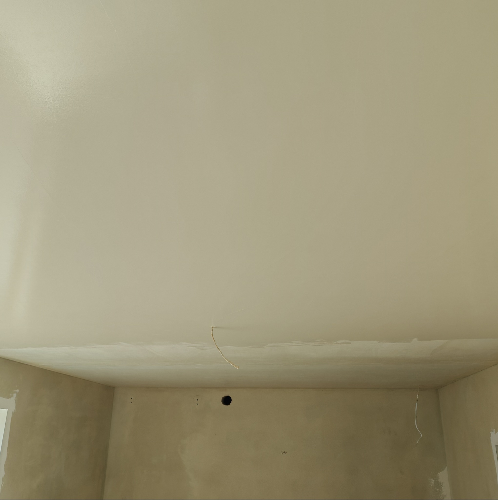 Wnętrze pokoju w trakcie prac wykończeniowych. Widoczny sufit po szpachlowaniu, ściany przygotowane do malowania. W tle widoczne otwory na instalacje.