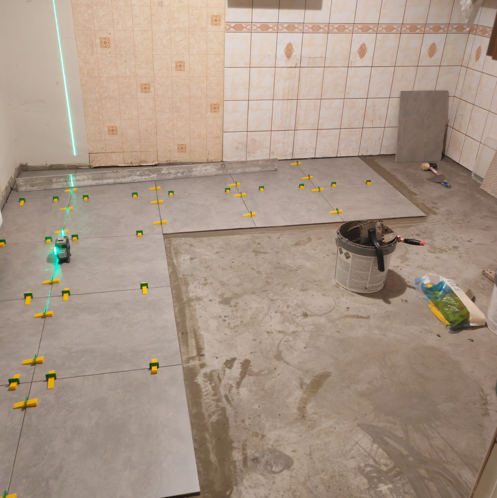 Układanie szarych płytek podłogowych w łazience z użyciem poziomicy laserowej i żółtych klinów dystansowych. Częściowo wykończone ściany z beżowych płytek.