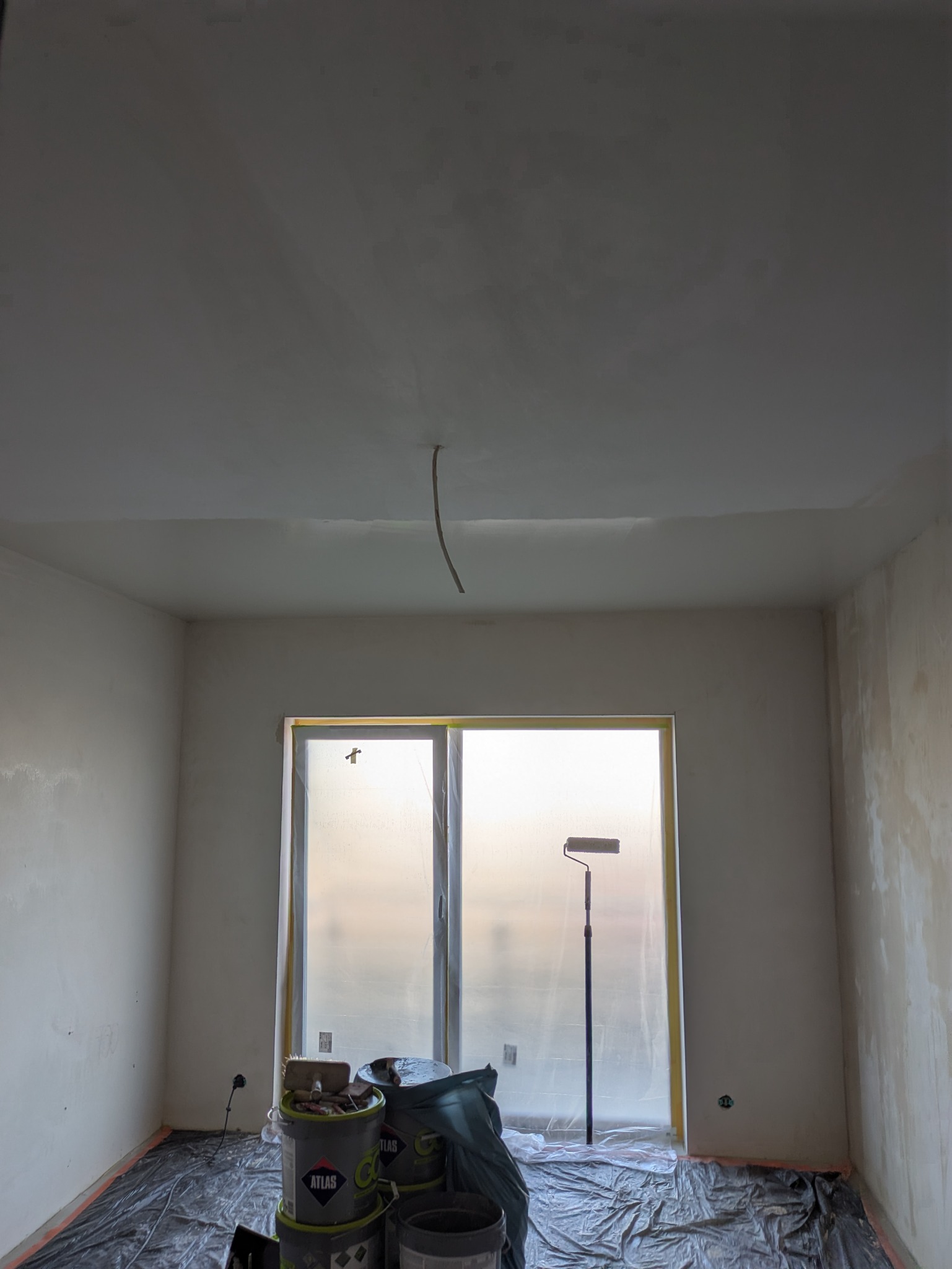 Pokój w trakcie malowania: ściany po szpachlowaniu, okno zabezpieczone folią, wałek malarski oparty o okno, wiaderka z farbą na podłodze przykrytej folią.