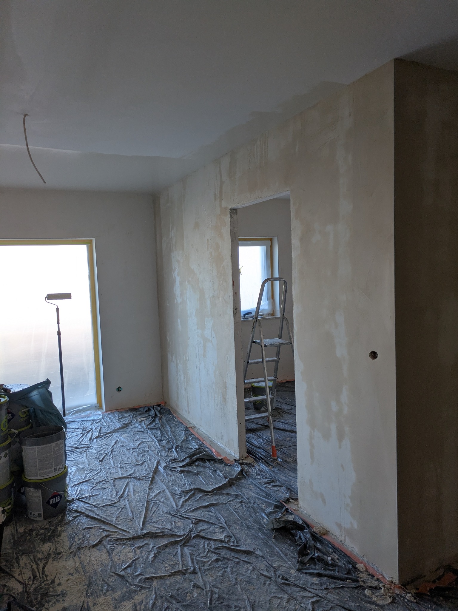 Pokój w trakcie szpachlowania: ściany pokryte gładzią, podłoga zabezpieczona folią, widoczna drabina i wałek malarski. Przygotowanie do malowania.