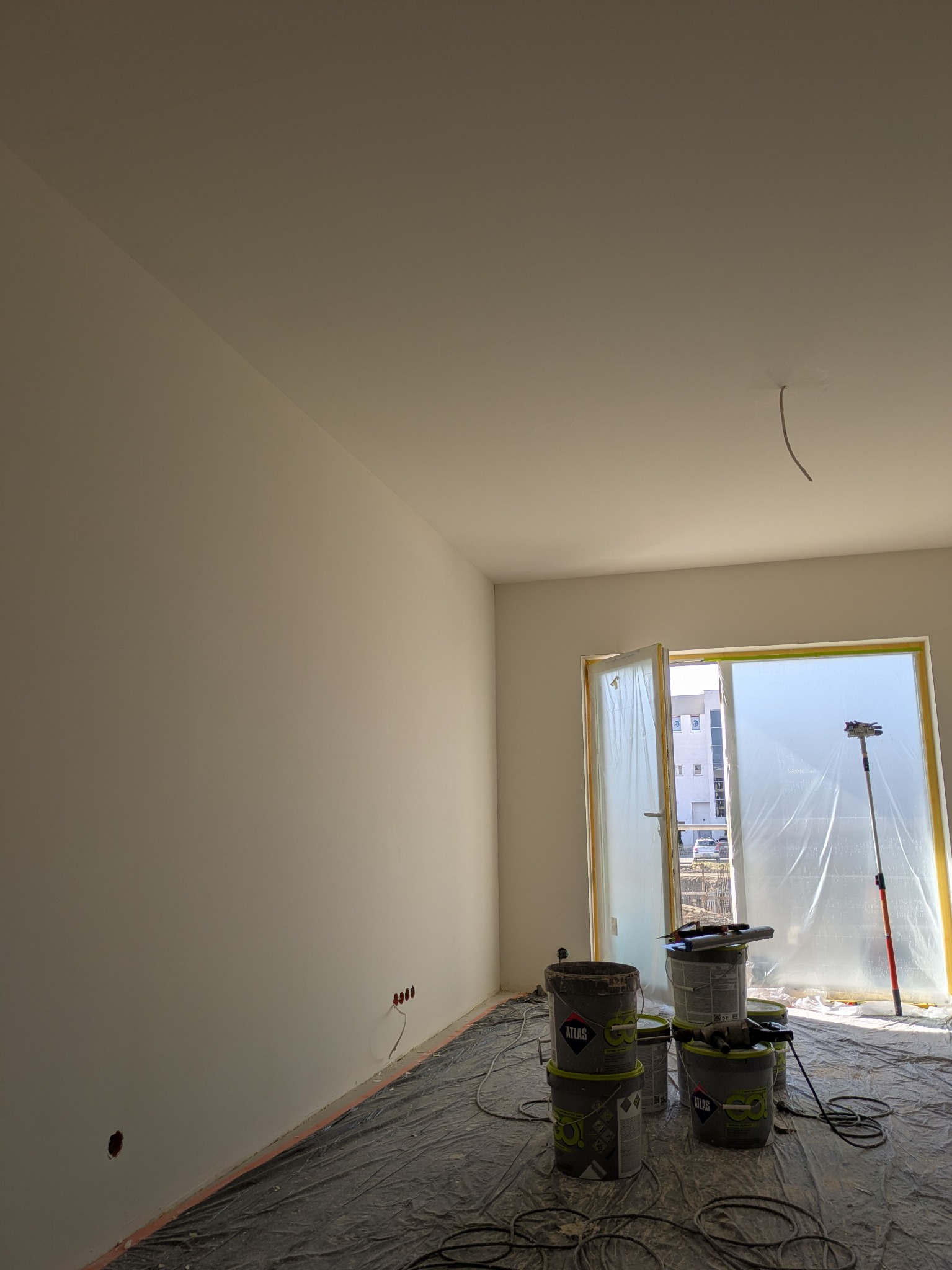 Wnętrze po świeżym malowaniu ścian na biało. Widoczne wiadra z farbą Atlas, zabezpieczona folią stolarka okienna i podłoga. Przygotowanie do dalszych prac wykończeniowych.