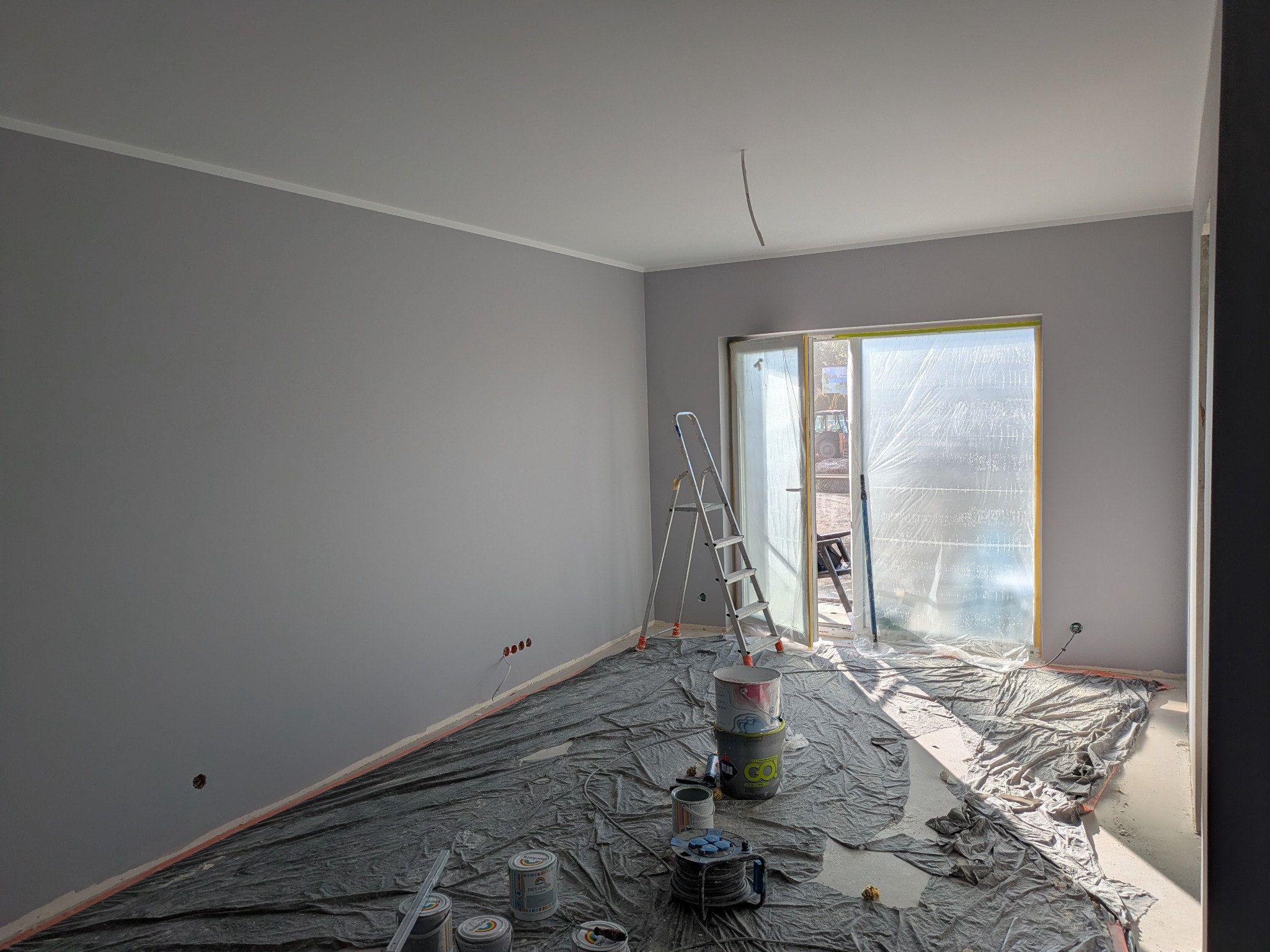 Pokój po malowaniu na szaro, zabezpieczony folią, z drabiną i puszkami farb. Widoczne gniazdka elektryczne i okno balkonowe osłonięte folią malarską.