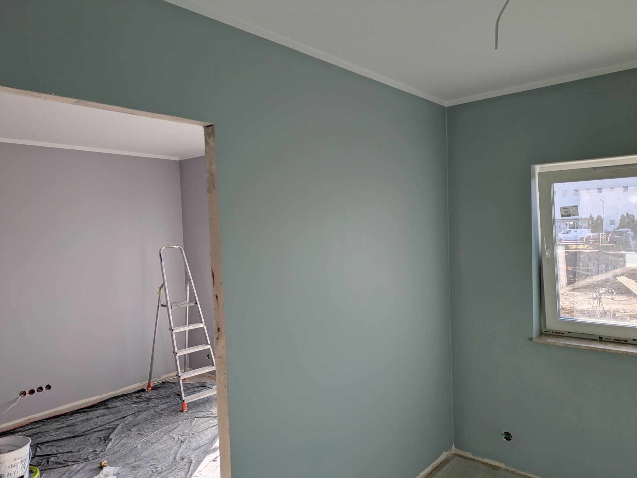 Świeżo pomalowane na gładko ściany w odcieniach szarości i błękitu, z widoczną drabiną i zabezpieczoną podłogą, ukazujące postęp prac wykończeniowych.