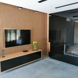 Inny Wymiar WKN - Nowoczesny salon z drewnianą ścianą z lameli, szafką RTV z jasnego drewna i czarnymi frontami, telewizorem, kominkiem i szarymi płytkami podłogowymi.