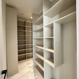 Pusta, jasna garderoba z półkami w zabudowie, marmurowa podłoga z wyraźnym wzorem żyłkowania, oświetlenie sufitowe punktowe.