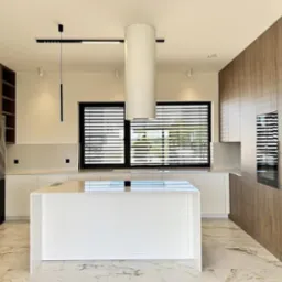 Nowoczesna kuchnia z wyspą, fronty szafek w odcieniach drewna, marmurowa podłoga, minimalistyczne oświetlenie i okno z żaluzjami.