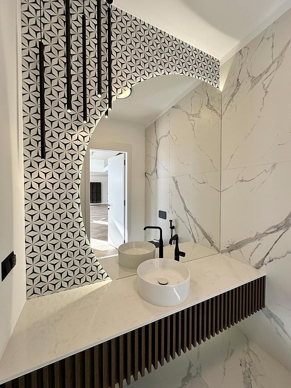 Nowoczesna łazienka z dwoma białymi umywalkami nablatowymi, czarną armaturą i lustrem w kształcie łuku, otoczonym wzorzystą mozaiką, oraz marmurowymi płytkami na ścianach.