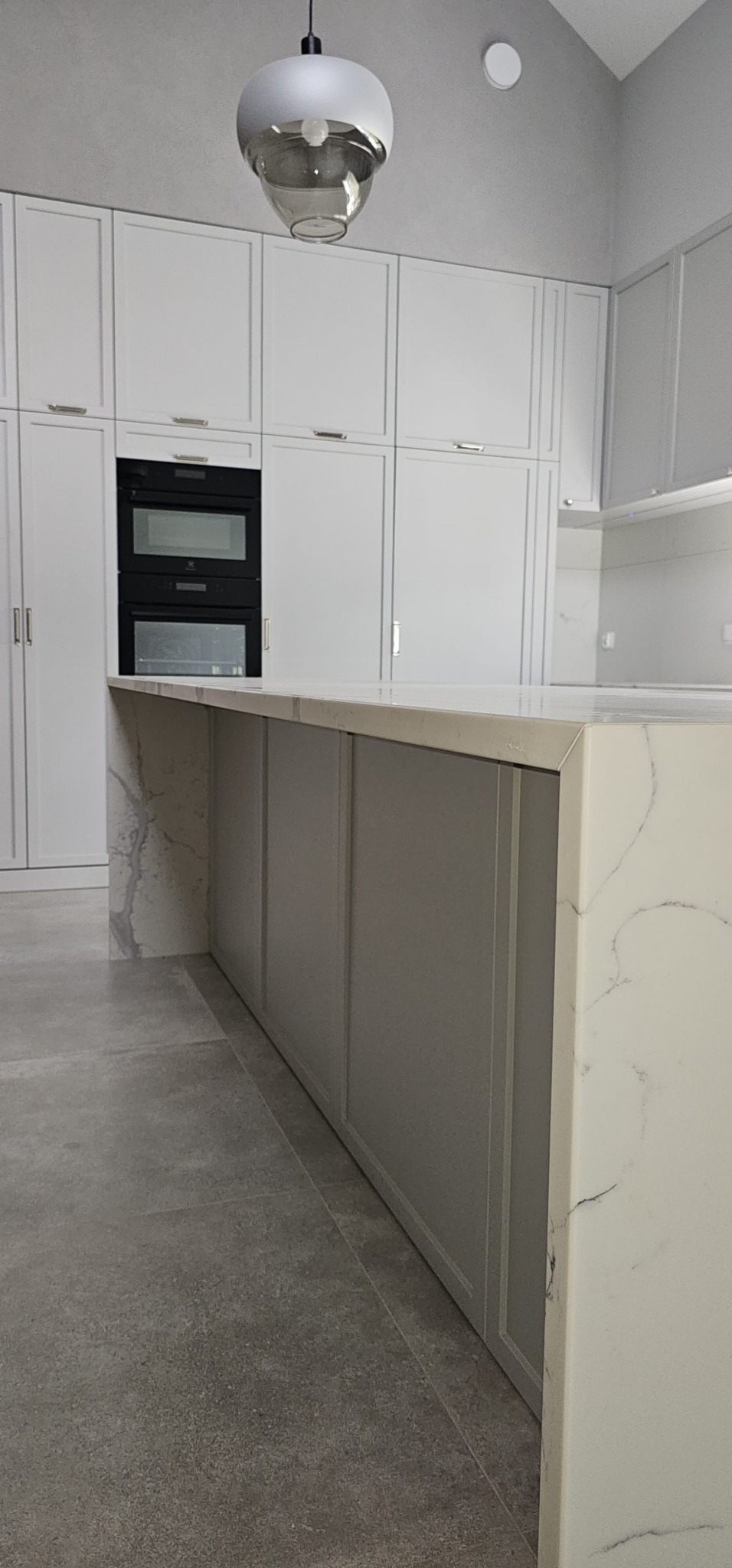 Jasna kuchnia z białymi szafkami, czarnym piekarnikiem w zabudowie i blatem z białego marmuru, oświetlona nowoczesną lampą wiszącą.