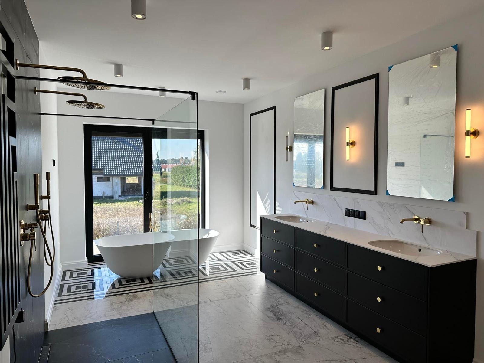 Nowoczesna łazienka z czarnymi szafkami pod umywalki z białym blatem imitującym marmur, trzema lustrami z oświetleniem, kabiną prysznicową typu walk-in i wolnostojącą wanną przy oknie z widokiem...