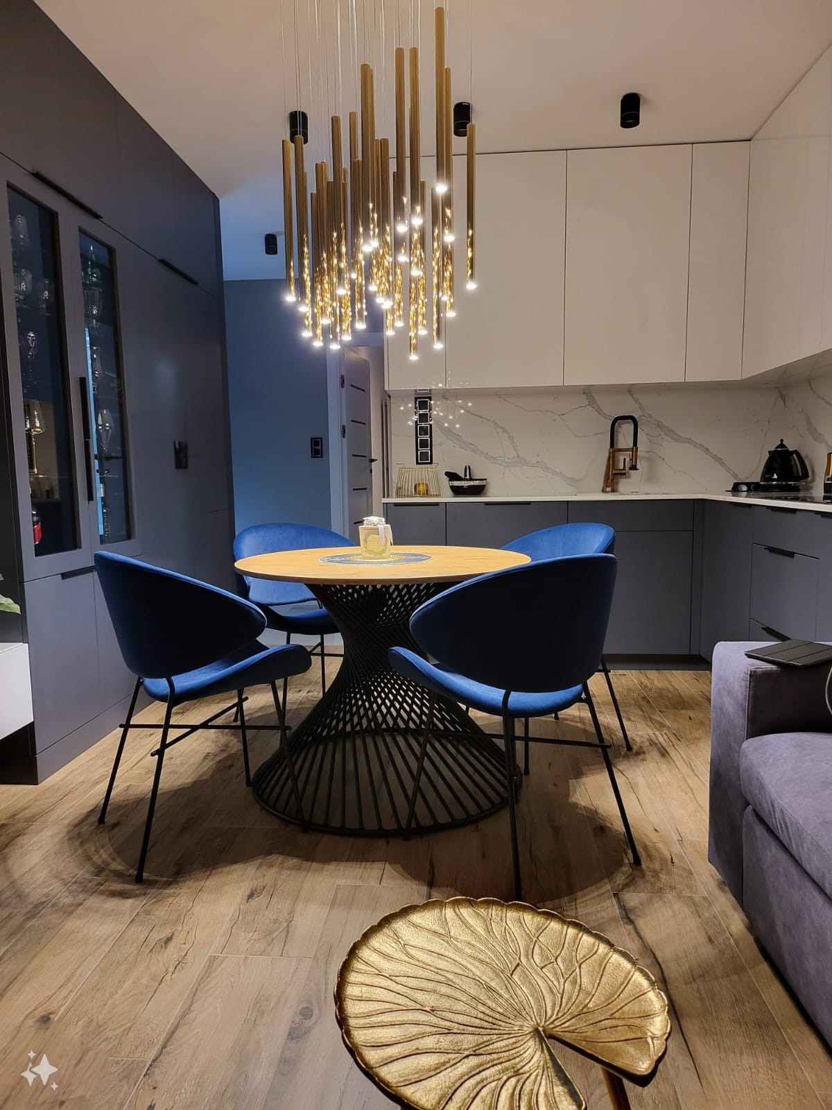 Nowoczesna aranżacja jadalni w Karbowie z okrągłym stołem na metalowej podstawie, niebieskimi krzesłami i designerską lampą wiszącą nad blatem, widok na zabudowę kuchenną z marmurowym panelem.