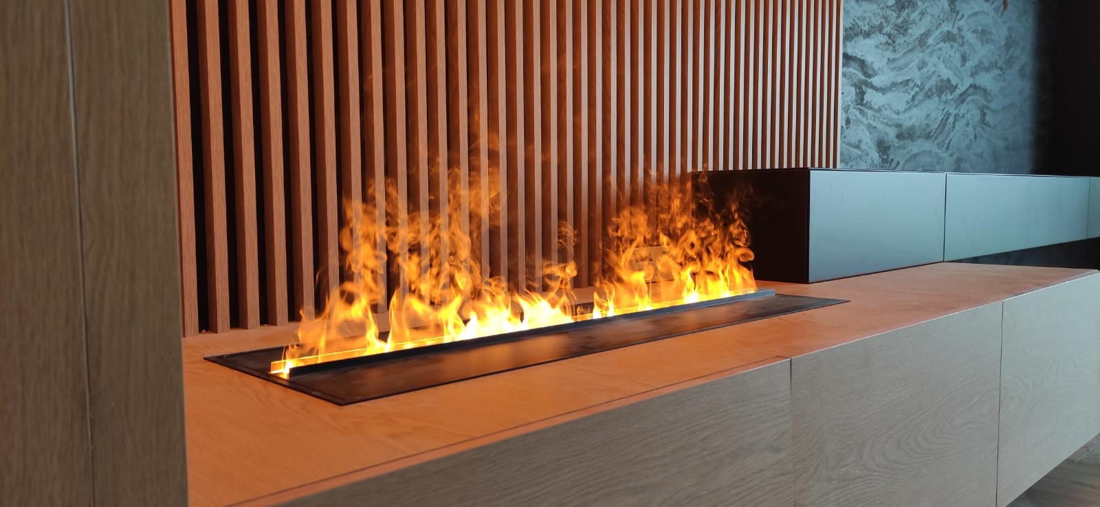 Nowoczesny biokominek wbudowany w drewnianą zabudowę meblową z pionowymi lamelami na ścianie, płomienie tańczące nad blatem, minimalistyczny design w odcieniach drewna i szarości.