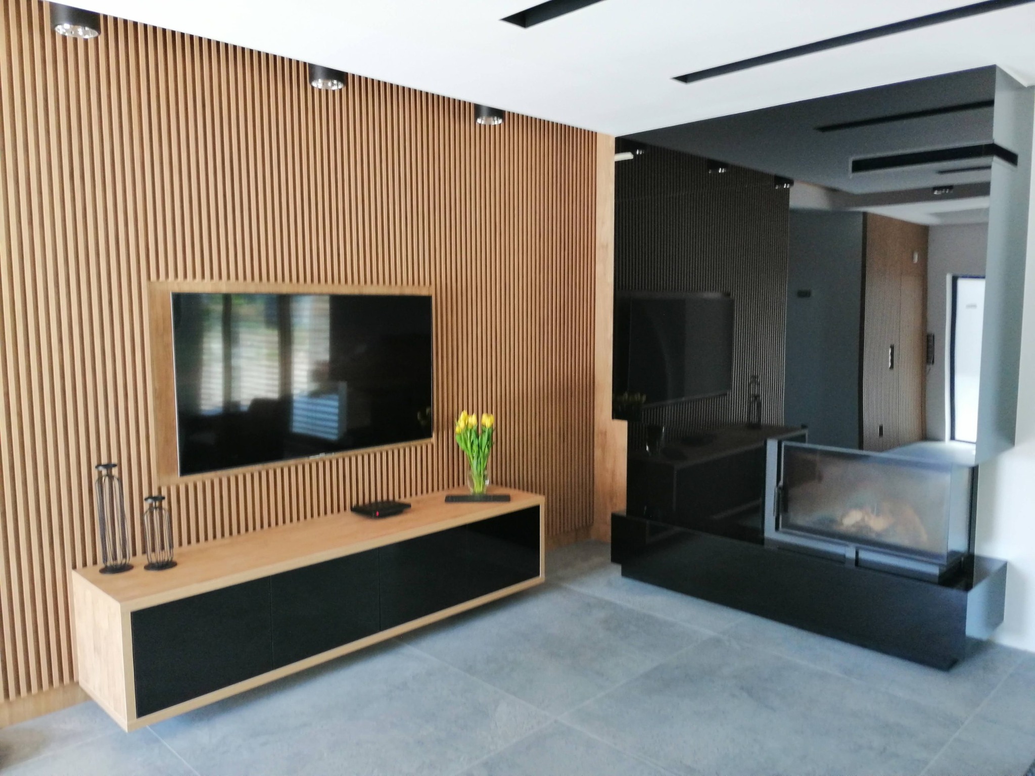 Nowoczesny salon z drewnianą ścianą z lameli, szafką RTV z jasnego drewna i czarnymi frontami, telewizorem, kominkiem i szarymi płytkami podłogowymi.