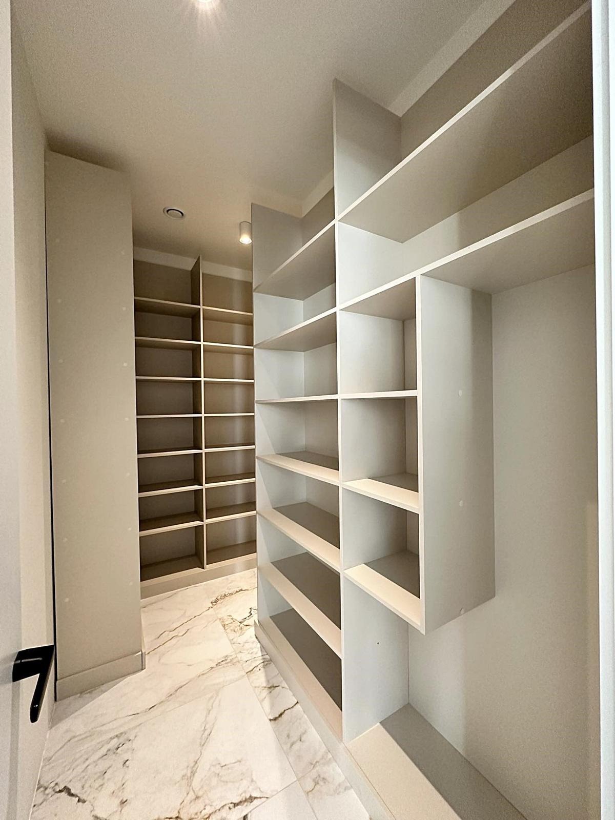 Pusta, jasna garderoba z półkami w zabudowie, marmurowa podłoga z wyraźnym wzorem żyłkowania, oświetlenie sufitowe punktowe.