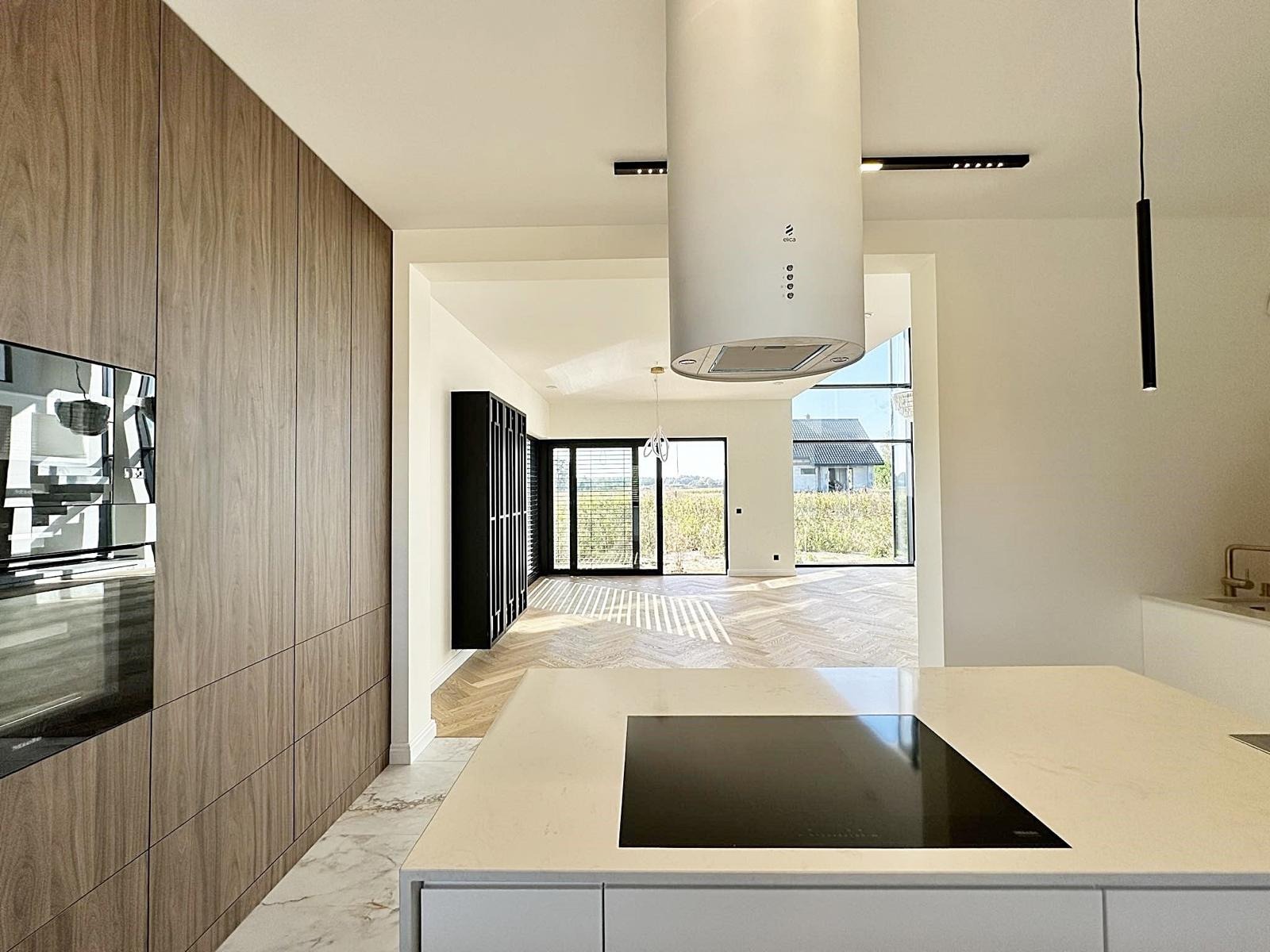 Nowoczesna kuchnia z drewnianą zabudową, białą wyspą kuchenną i cylindrycznym okapem, parkiet w jodełkę na podłodze, widok na zewnątrz przez duże okna.