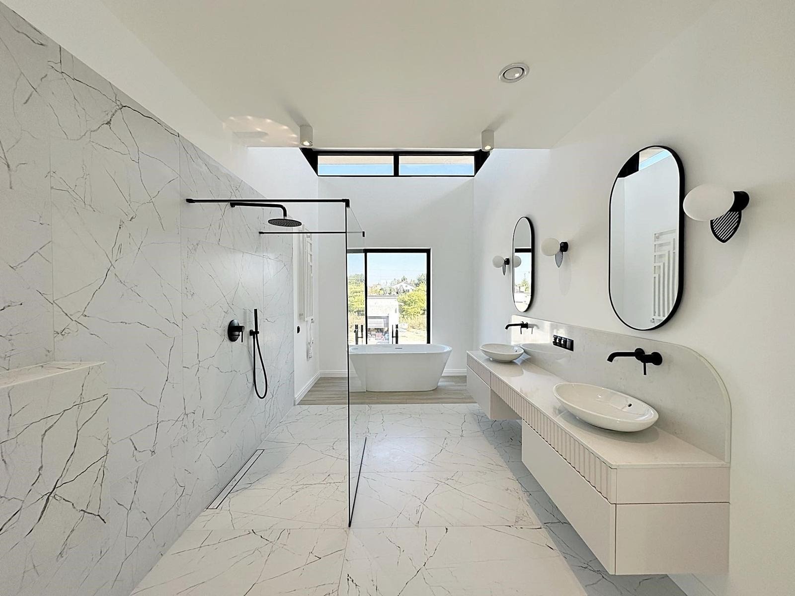 Nowoczesna łazienka z marmurowymi płytkami, kabiną prysznicową typu walk-in z czarną armaturą, podwójną umywalką nablatową na szafce z ryflowanym frontem oraz owalnymi lustrami z oświetleniem.