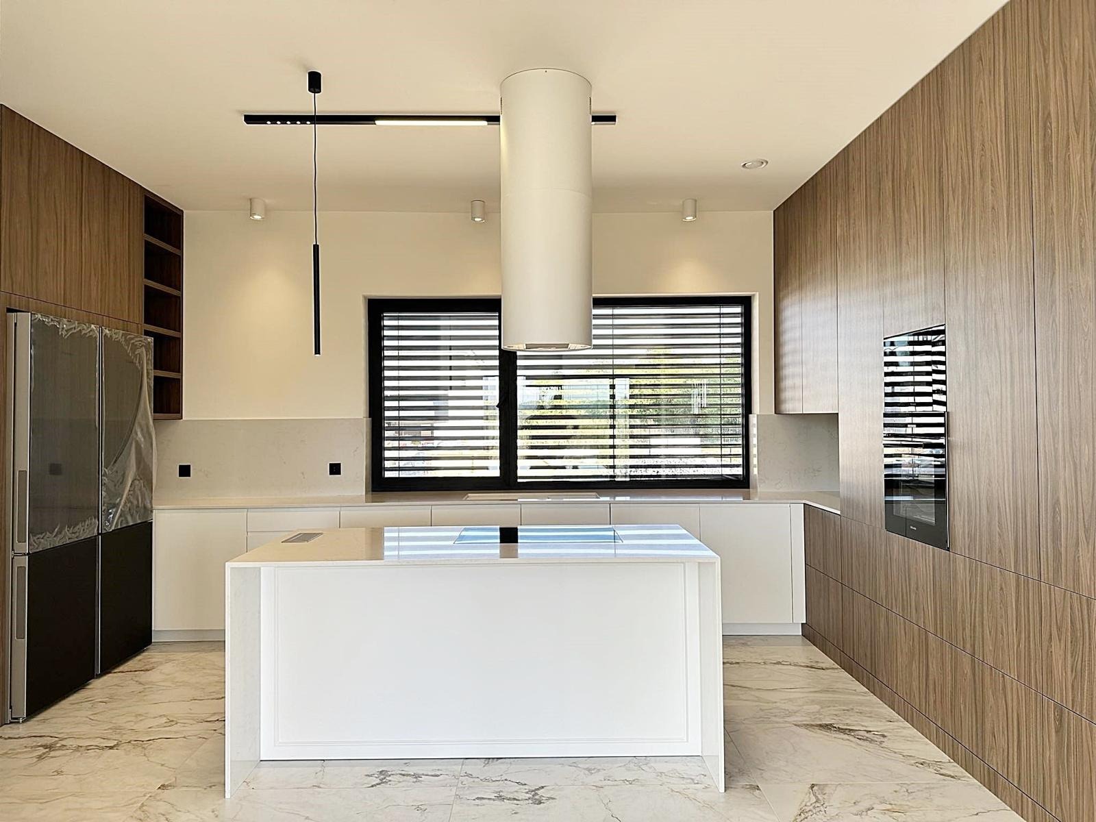 Nowoczesna kuchnia z wyspą, fronty szafek w odcieniach drewna, marmurowa podłoga, minimalistyczne oświetlenie i okno z żaluzjami.